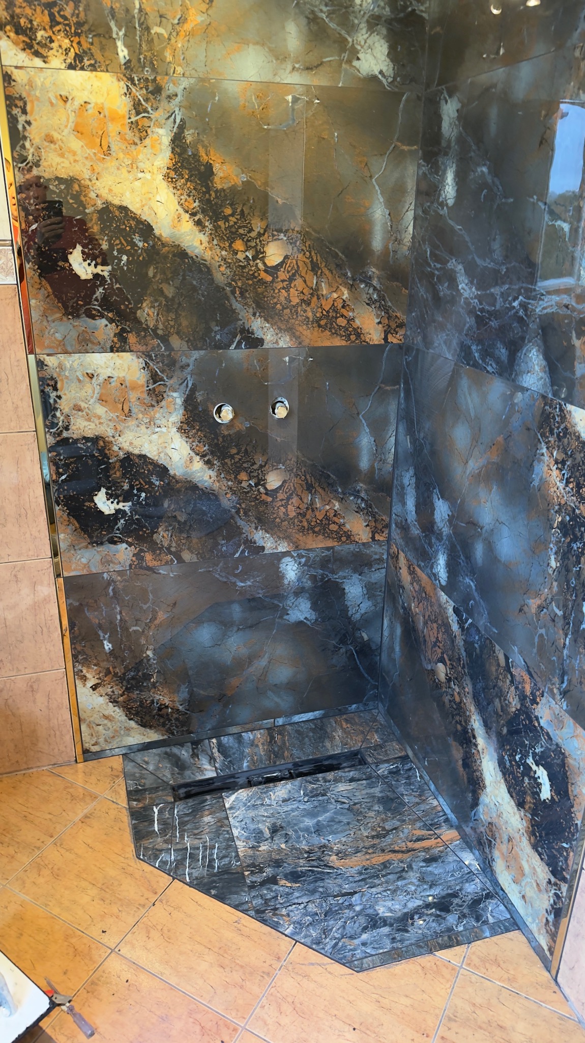 Wykończona kabina prysznicowa z ciemnymi płytkami imitującymi marmur, widoczny odpływ liniowy i przygotowane otwory na armaturę. Ułożona podłoga z płytek w odcieniach beżu.