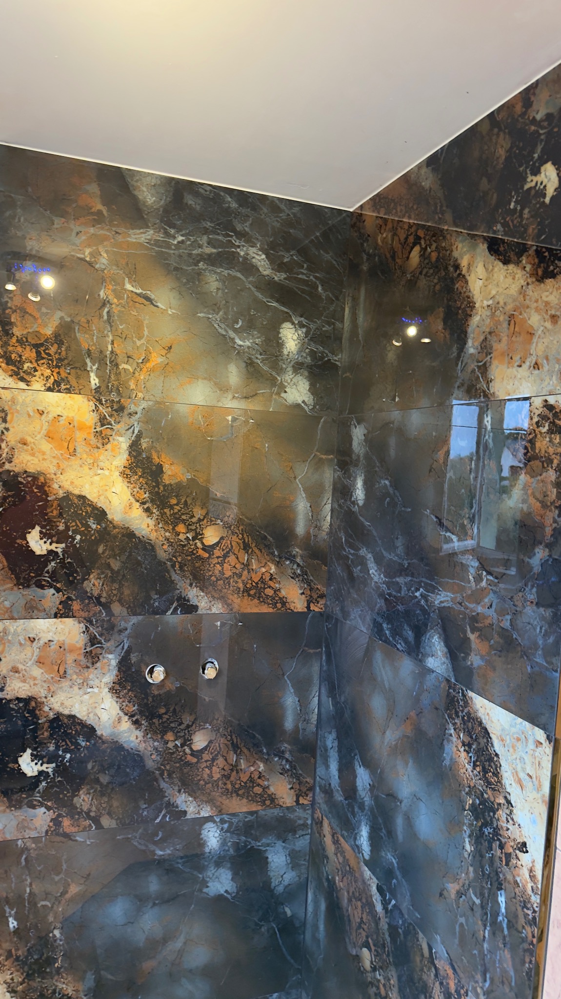 Wykończona łazienka z luksusowymi, wielkoformatowymi płytkami imitującymi marmur w odcieniach brązu i szarości. Widoczne punkty świetlne i fragment armatury.