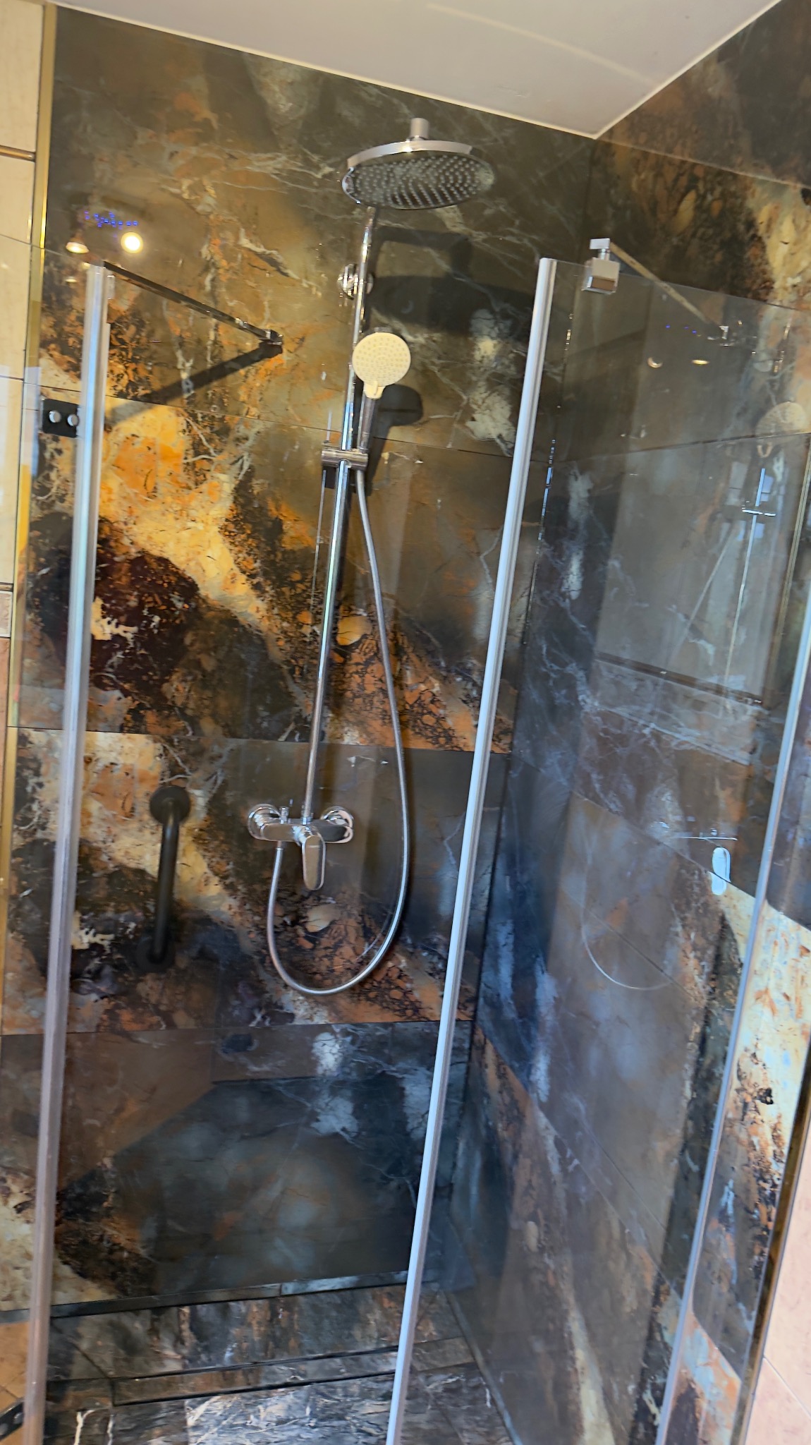 Kabina prysznicowa z deszczownicą i szklanymi drzwiami na tle ściany wyłożonej płytkami imitującymi marmur w odcieniach brązu i szarości.