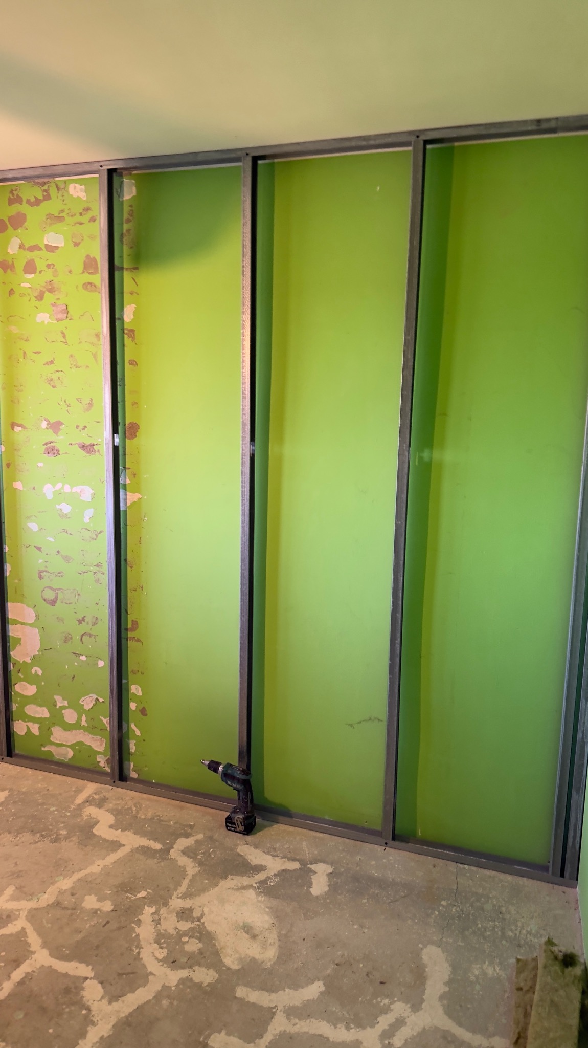 Montaż stelaża z profili metalowych do płyty G-K na ścianie w odcieniach zieleni. W tle widoczny fragment ściany z resztkami farby. Na podłodze leży wkrętarka.