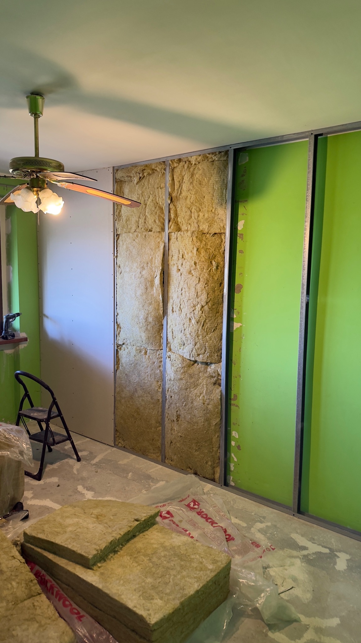 Montaż izolacji z wełny mineralnej w metalowym stelażu na ścianie. Widoczne płyty gipsowo-kartonowe i narzędzia. Etap prac wykończeniowych w pomieszczeniu z zielonymi ścianami.