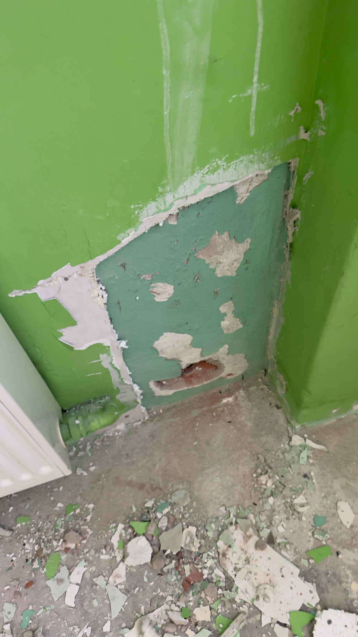 Fragment ściany w jaskrawej zieleni z uszkodzeniami i odpadającym tynkiem, ukazujący warstwy farby i surowy mur. Na podłodze widoczne odłamki i resztki materiałów budowlanych.