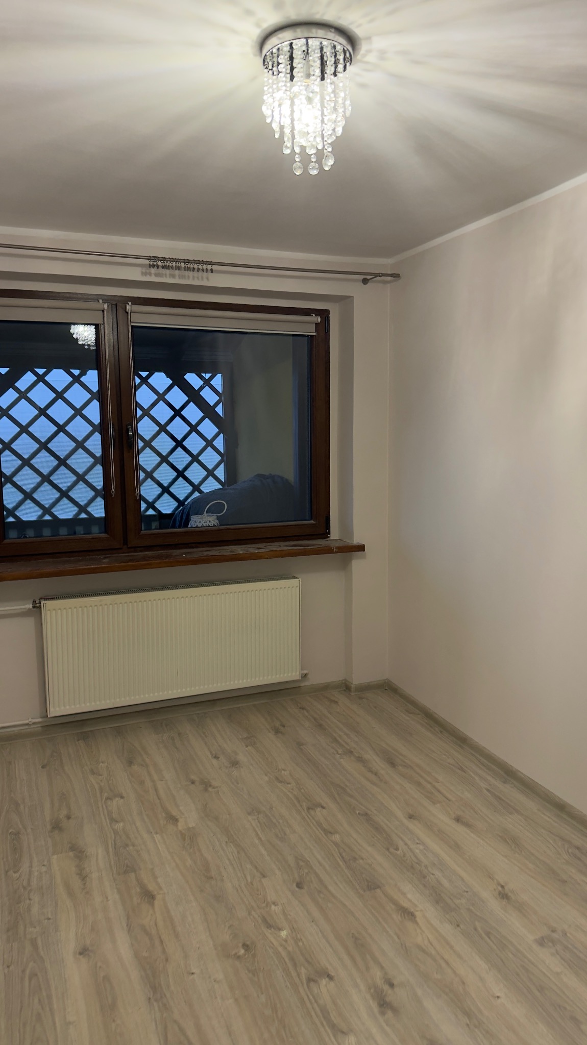 Jasne wnętrze pokoju po remoncie: panele podłogowe, okno z kratą, grzejnik i ozdobny żyrandol. Ściany pomalowane na jasny odcień.