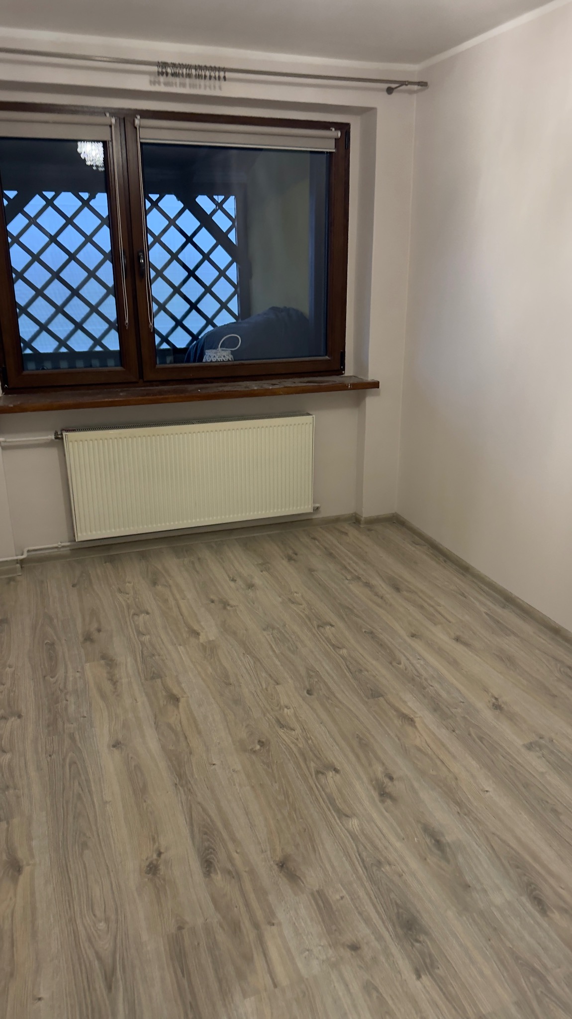 Jasne panele podłogowe w pokoju z brązowym oknem z kratką i białym grzejnikiem. Ściany pomalowane na jasny odcień bieli. Widok na fragment pokoju po remoncie.