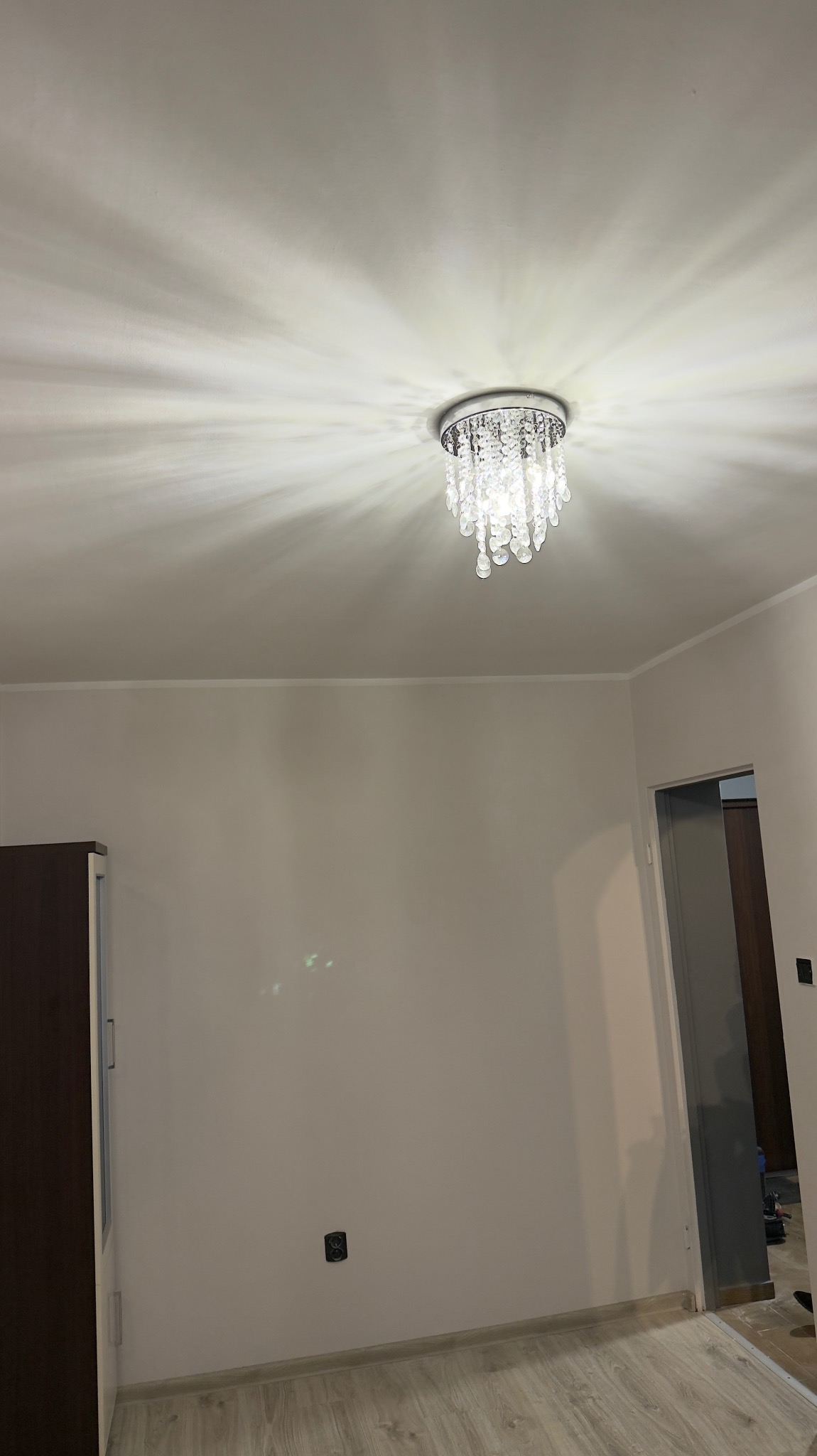 Jasne wnętrze po odświeżeniu: kremowe ściany, parkiet, ozdobny żyrandol z kryształkami. Widoczny fragment szafy i drzwi. Gniazdko elektryczne na ścianie.