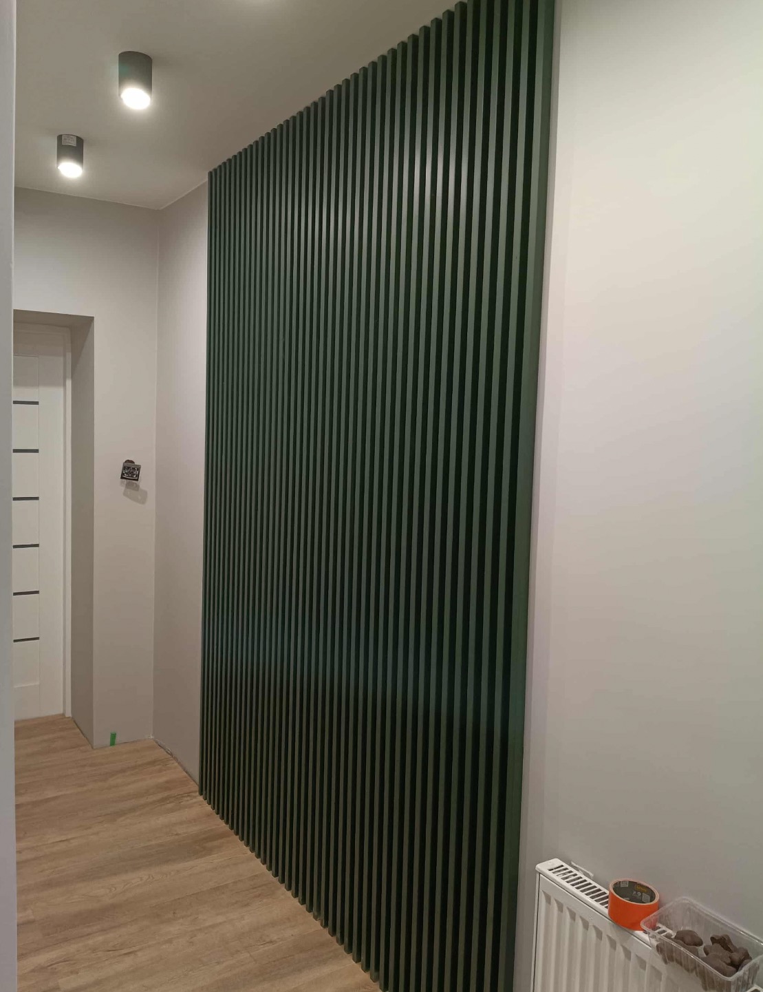 Zielone lamele dekoracyjne na ścianie w korytarzu, widoczne oświetlenie sufitowe i fragment grzejnika. Podłoga wyłożona panelami. Nowoczesny design.