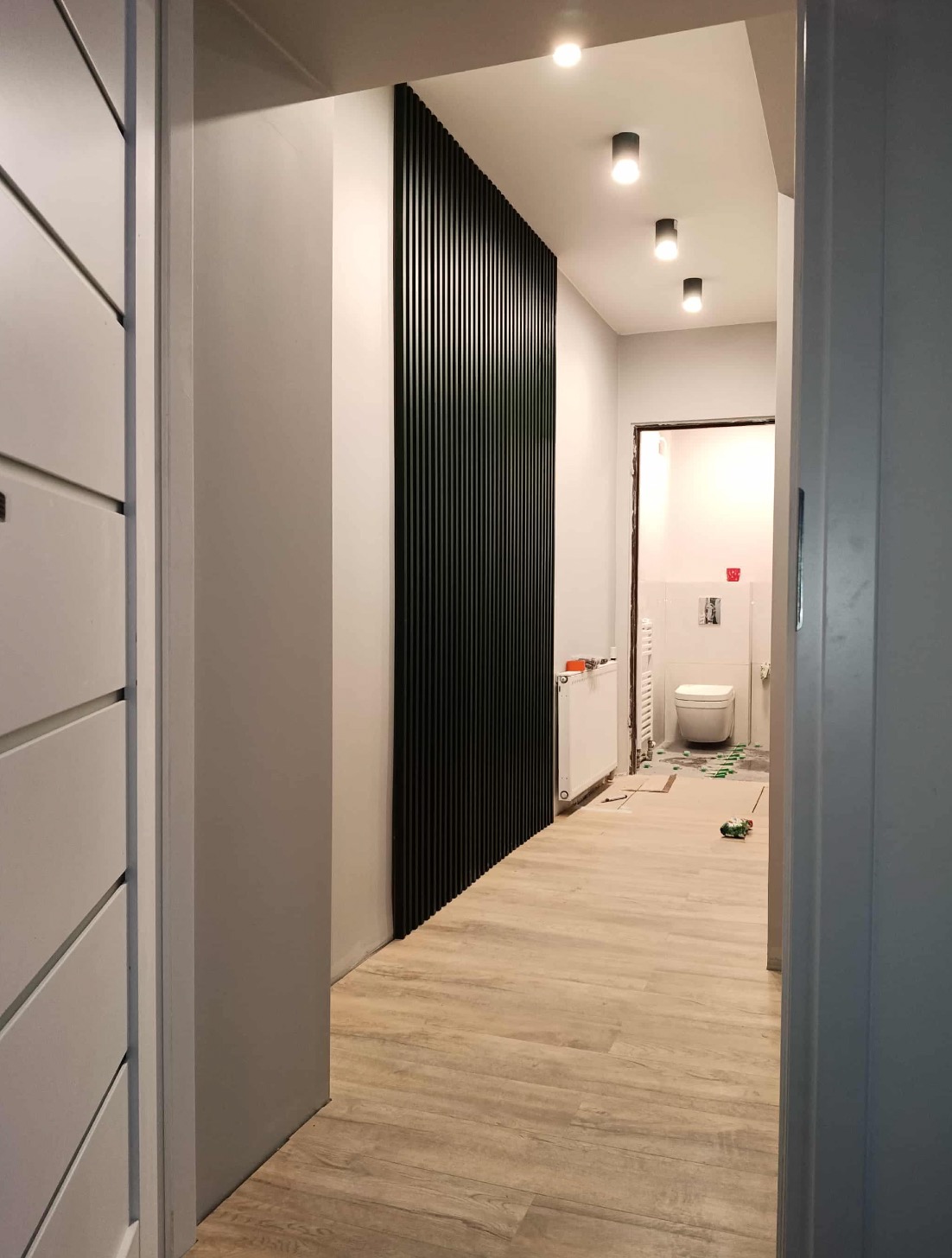 Nowoczesny korytarz z panelami ściennymi, jasną podłogą i oświetleniem punktowym. Widok na łazienkę w trakcie wykończenia.