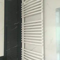 Biały, łazienkowy grzejnik drabinkowy na tle ściany z czarno-białego marmuru, widoczny fragment kabiny prysznicowej.