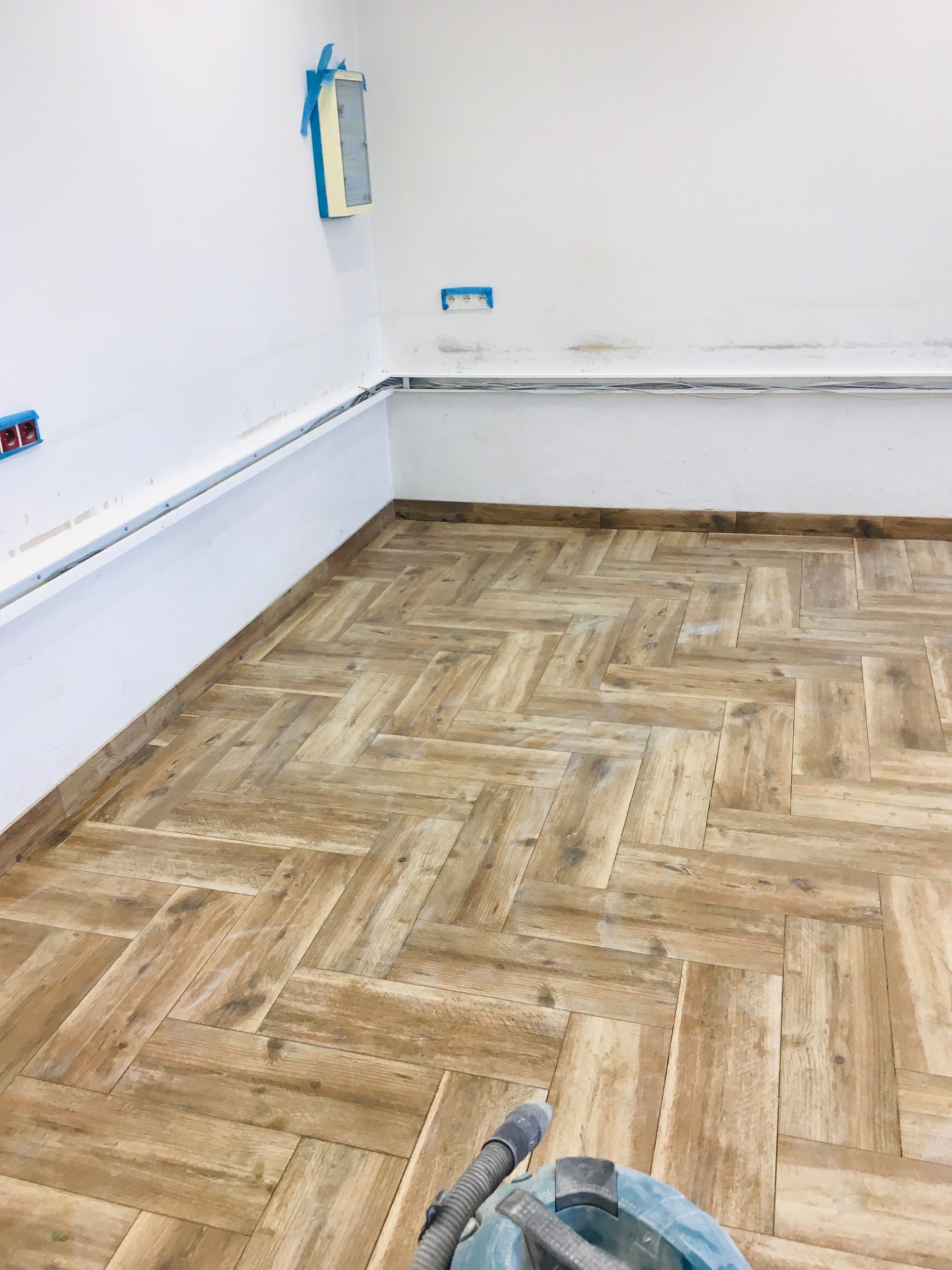 Ułożona, drewnopodobna glazura w jodełkę na podłodze, widoczny odkurzacz przemysłowy, ściany przygotowane do malowania, zabezpieczone gniazdka i listwy przypodłogowe.
