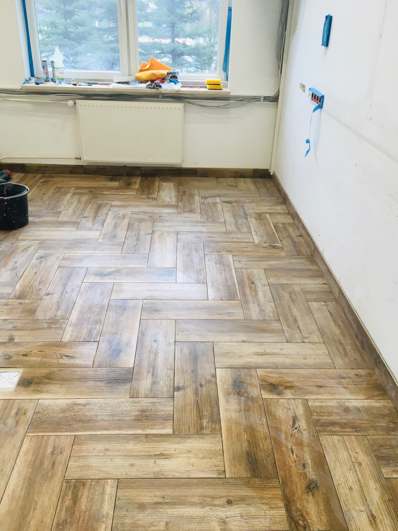 Ułożona podłoga z płytek imitujących drewno w jodełkę, widoczne przygotowane gniazdka elektryczne i okno z widokiem na drzewa.