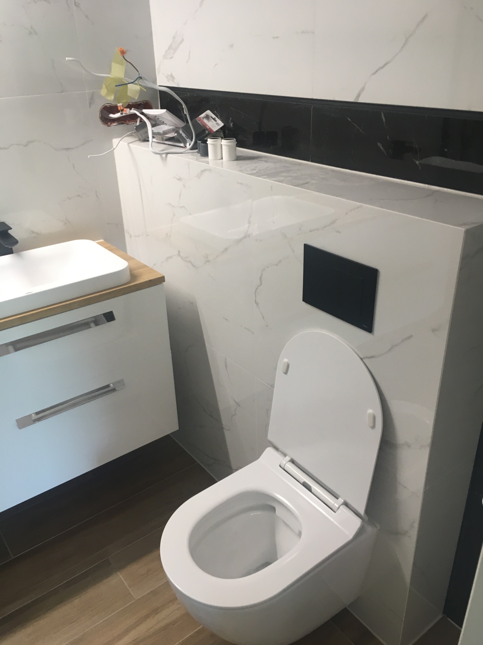 Wnętrze łazienki w trakcie prac wykończeniowych: biała toaleta wisząca z otwartą klapą, umywalka z szafką i drewnianym blatem, marmurowe płytki na ścianach, widoczne kable i materiały budowlane...