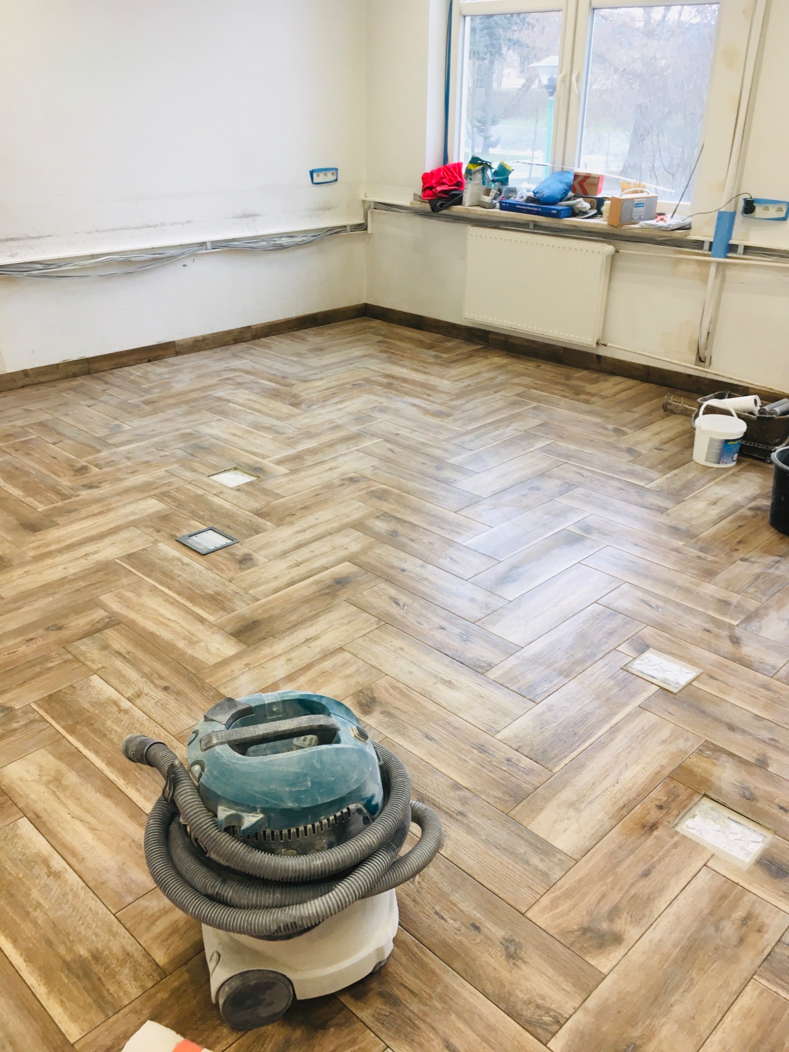 Ułożona w jodełkę podłoga z płytek imitujących drewno, z widocznym odkurzaczem przemysłowym, niezamaskowanymi otworami rewizyjnymi i akcesoriami malarskimi na parapecie okna.