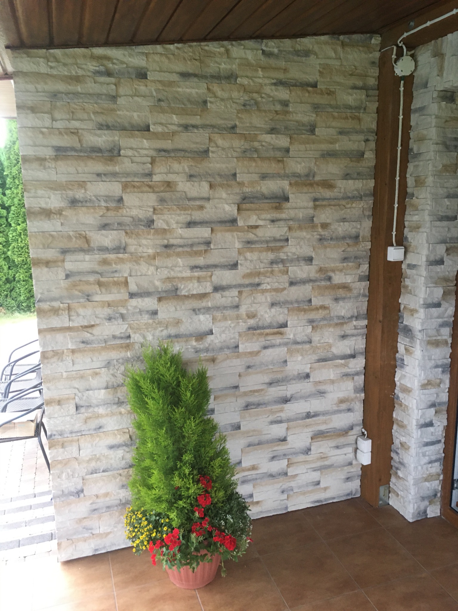 Ściana zewnętrzna wyłożona dekoracyjną okładziną imitującą kamień, z widoczną instalacją elektryczną i donicą z iglakiem oraz kwiatami.