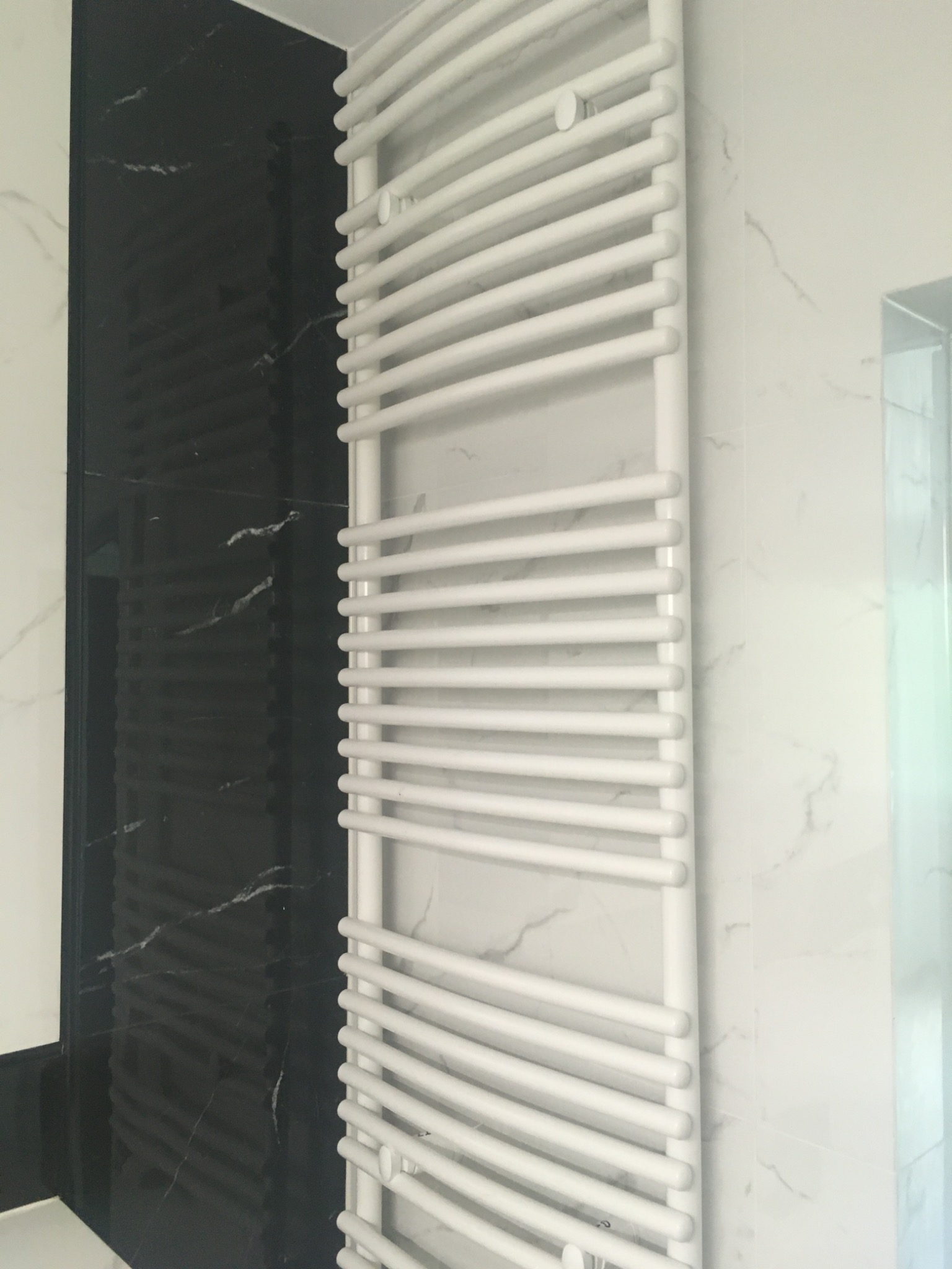 Biały, łazienkowy grzejnik drabinkowy na tle ściany z czarno-białego marmuru, widoczny fragment kabiny prysznicowej.
