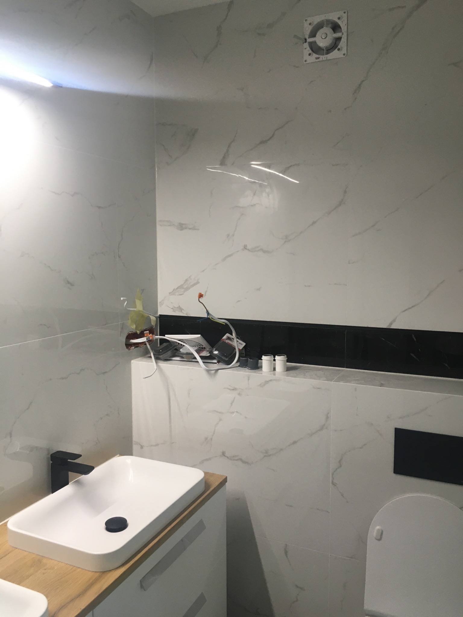 Łazienka w trakcie remontu: białe płytki imitujące marmur, czarny pas płytek, umywalka na drewnianym blacie, widoczne przewody elektryczne i puszka instalacyjna bez osprzętu, wentylator sufitowy...