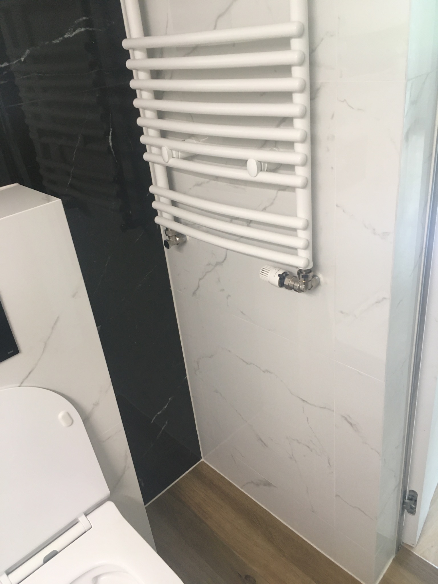 Biały grzejnik drabinkowy zamontowany na ścianie z białych płytek imitujących marmur w nowoczesnej łazience z czarnymi akcentami, widoczny fragment toalety i drewnopodobnej podłogi.