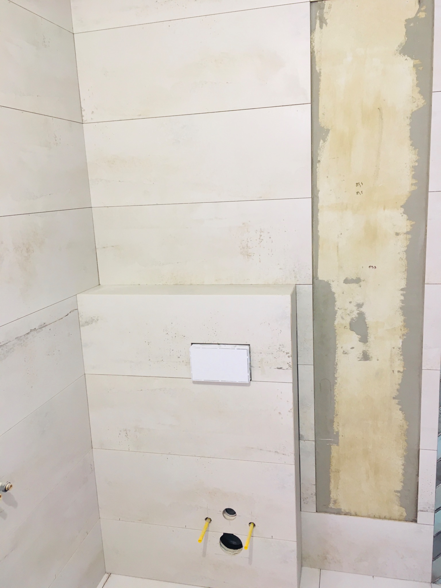 Ściana łazienki w trakcie prac wykończeniowych: częściowo ułożone białe, poziome płytki imitujące drewno, widoczne instalacje wodno-kanalizacyjne oraz fragment ściany przygotowany pod płytki...