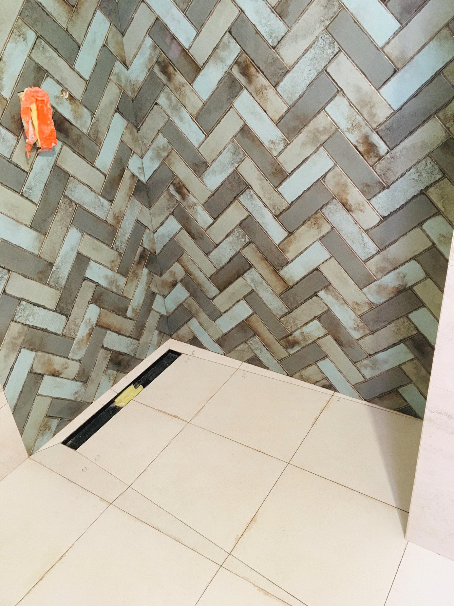 Ściana łazienki wyłożona mozaiką z podłużnych płytek w odcieniach szarości, błękitu i beżu, ułożonych w jodełkę; widoczny fragment podłogi z jasnych, kwadratowych płytek i czarny odpływ liniowy.