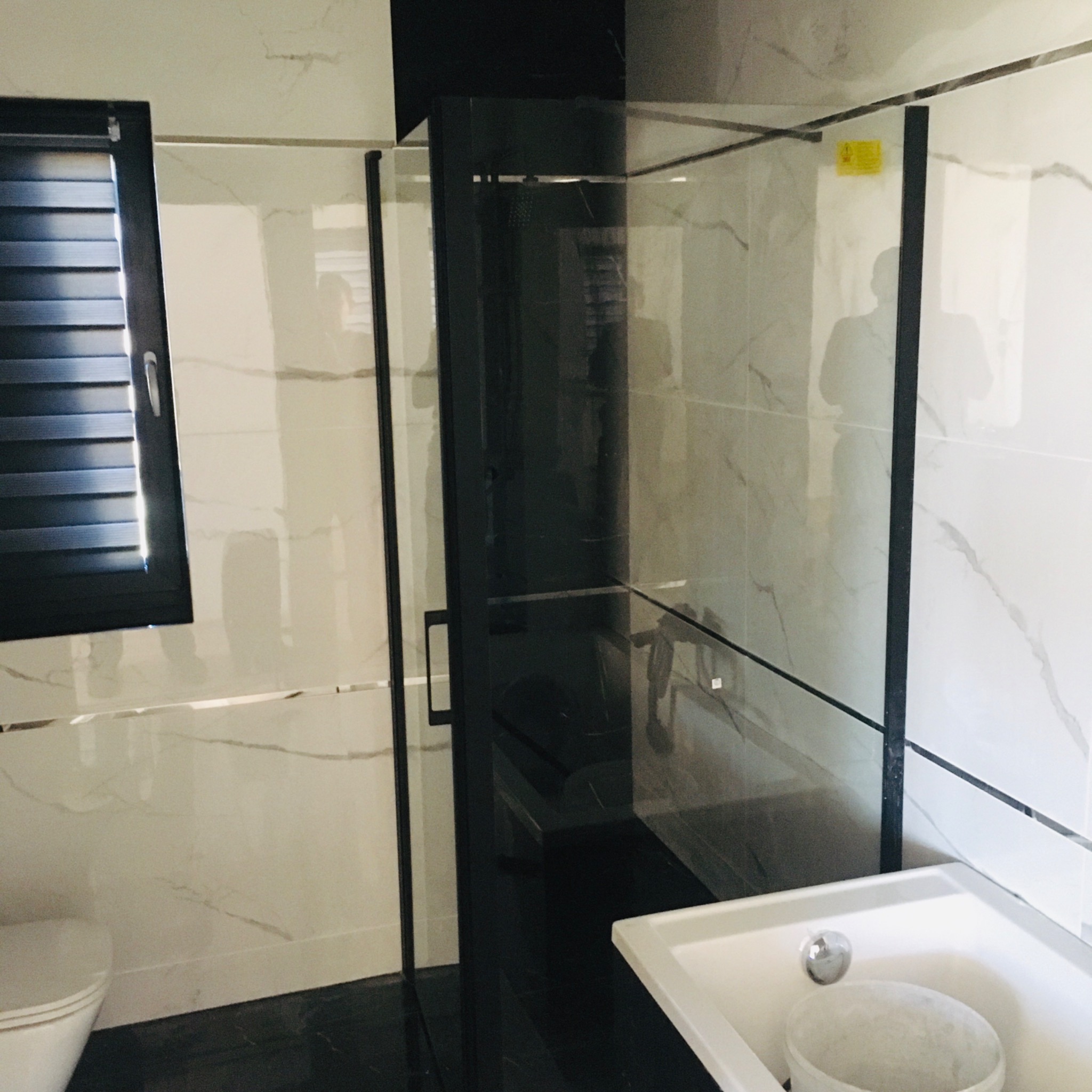 Wykończona łazienka z kabiną prysznicową z czarnymi ramami, białą umywalką i marmurowymi płytkami na ścianach, widoczne czarne żaluzje w oknie.