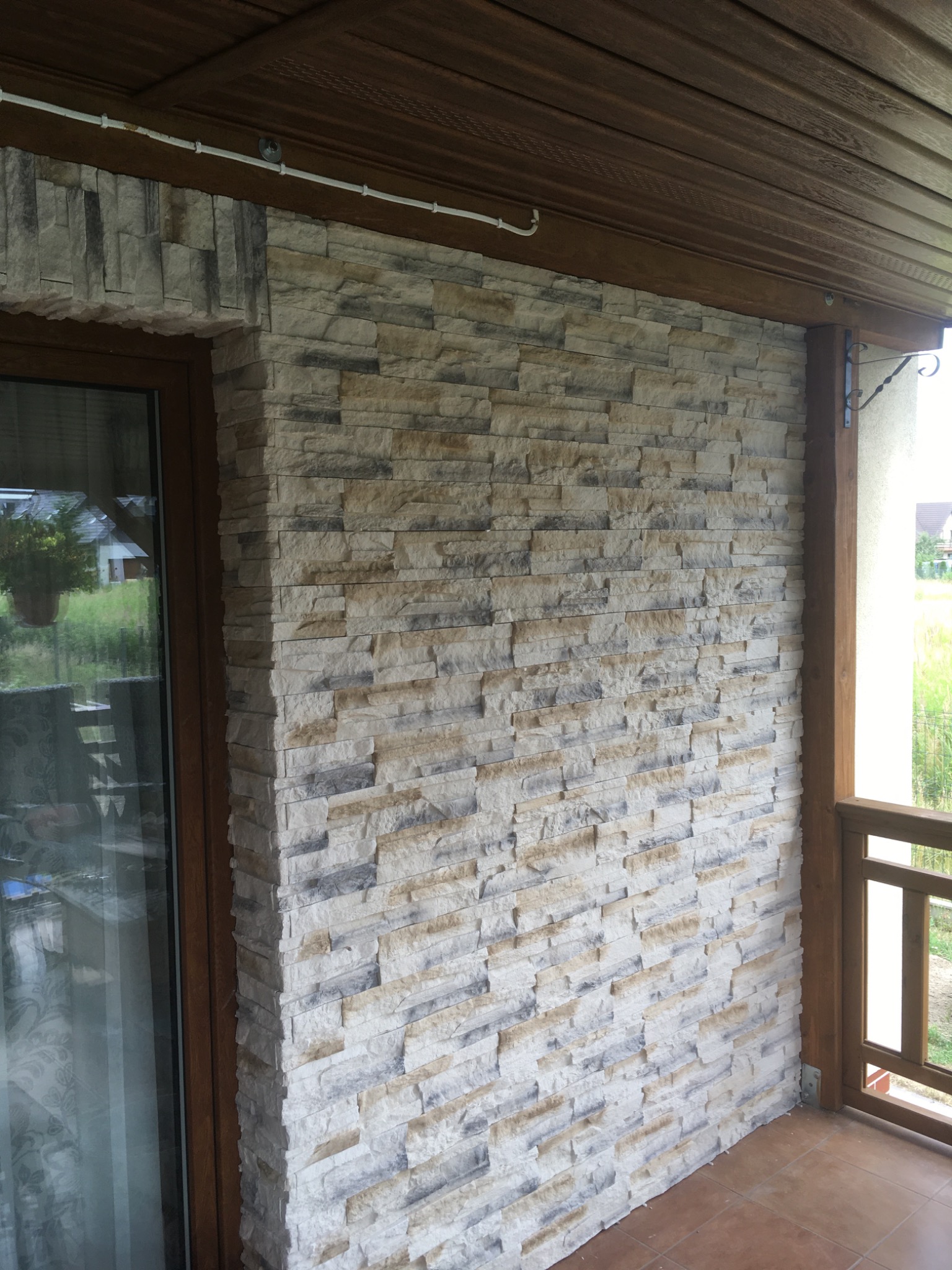 Ściana zewnętrzna wykończona dekoracyjnym kamieniem elewacyjnym w odcieniach beżu i szarości, z widocznym oknem z brązową ramą i fragmentem drewnianego podbitki dachowej.