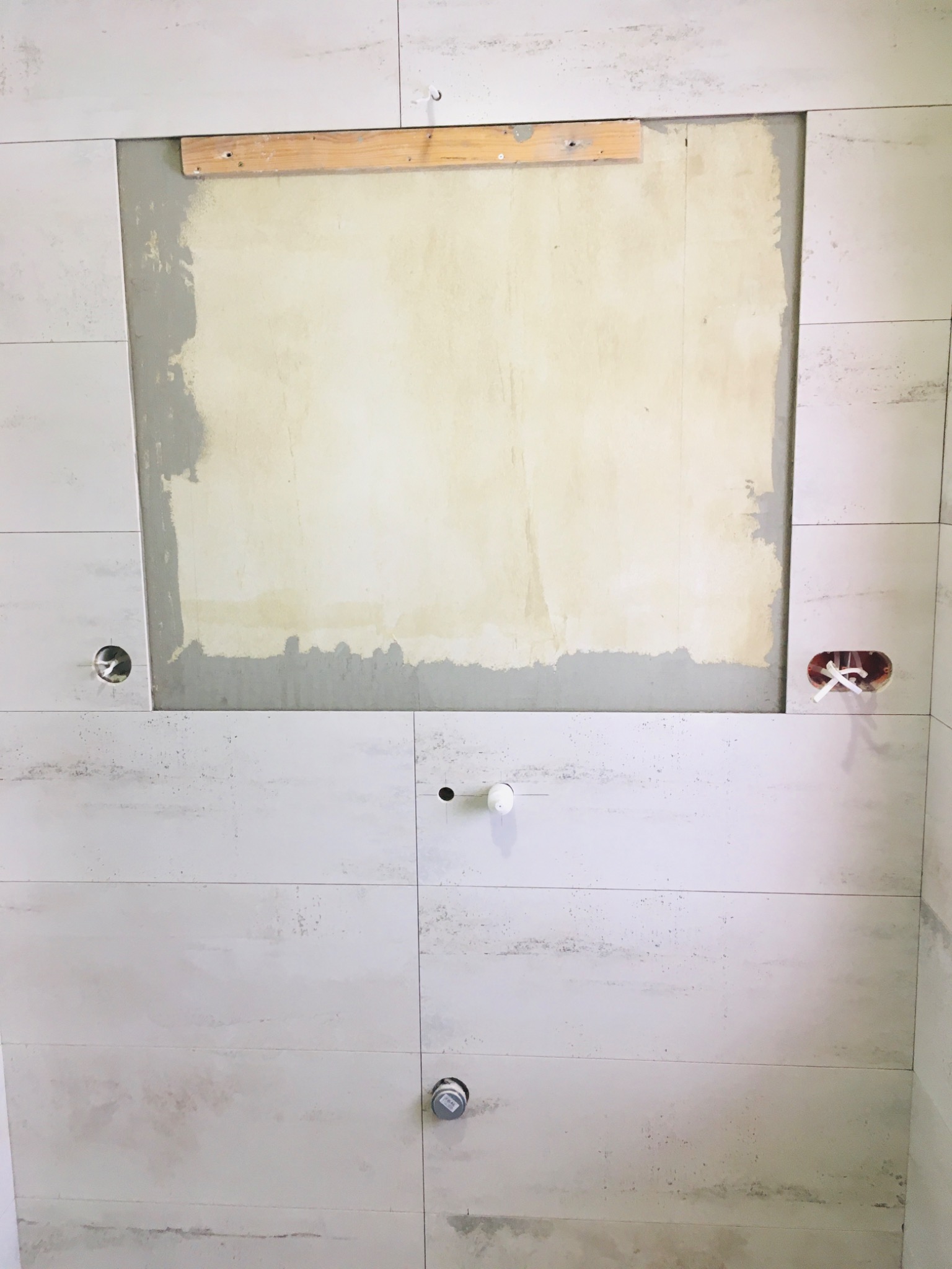 Ściana łazienki w trakcie układania płytek imitujących kamień, z przygotowanym miejscem na lustro lub szafkę i widocznymi otworami na instalacje.