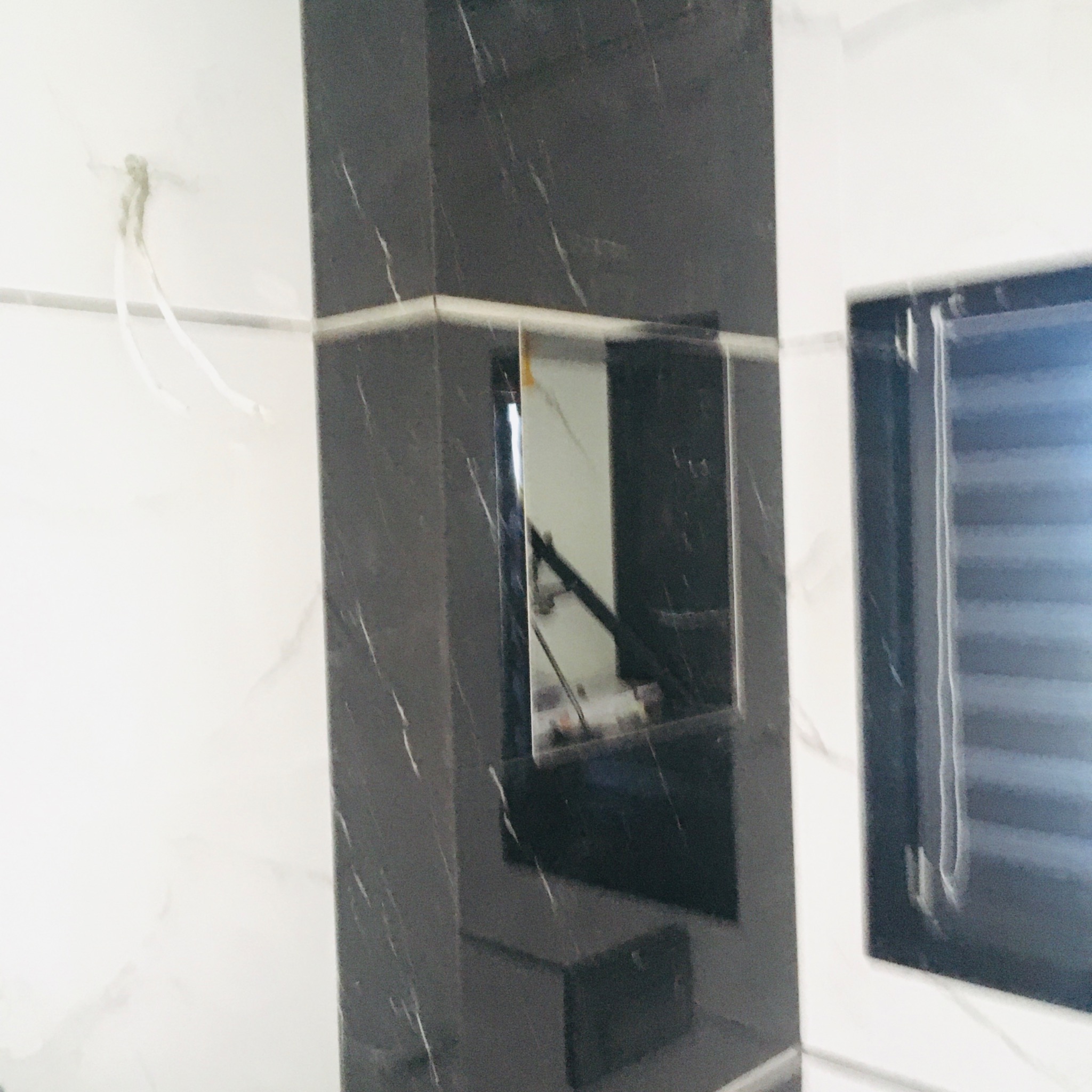 Fragment łazienki z wykończeniem w czarnym i białym marmurze, widoczne lustro wbudowane w czarną ścianę oraz fragment okna z żaluzjami, w tle białe ściany z delikatnym wzorem marmuru.