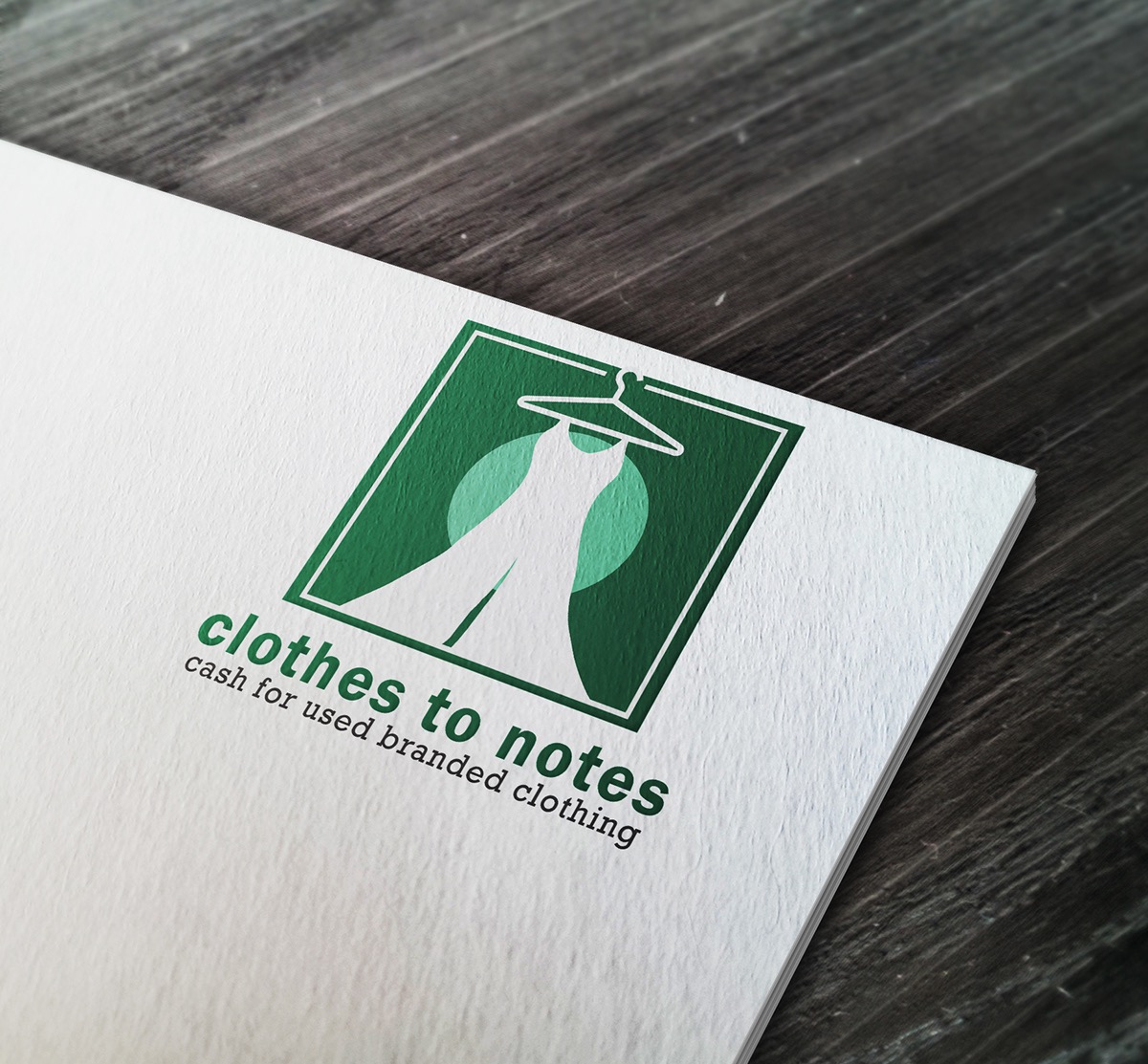 Logo firmy odzieżowej 'clothes to notes' na białym papierze z teksturą, na tle ciemnego drewnianego stołu. Logo przedstawia sukienkę wiszącą na wieszaku wewnątrz zielonego kwadratu z jasnym...