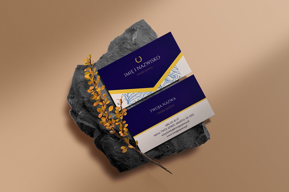 Dwie wizytówki z granatowo-białym motywem, ułożone na szarym kamieniu z gałązką z żółtymi liśćmi na beżowym tle.