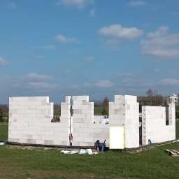 Grzegorz Kozieł - Wznoszenie ścian parteru domu z białych bloczków, widok z zewnątrz na tle zielonej łąki i błękitnego nieba z chmurami. Widoczne narzędzia i materiały budowlane.