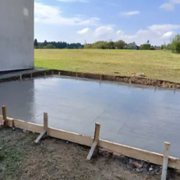 Świeżo wylany, gładki fundament pod budynek z drewnianym szalunkiem, na tle zielonego pola i błękitnego nieba z chmurami. Widok z poziomu gruntu.