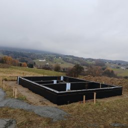 Grzegorz Kozieł - Zabezpieczone fundamenty domu jednorodzinnego na tle pagórkowatego krajobrazu w pochmurny dzień. Widoczne zbrojenia i izolacja termiczna.