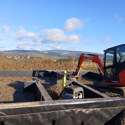Grzegorz Kozieł - Fundamenty domu w trakcie budowy: pomarańczowa koparka Kubota, ubijarka, czarna folia izolacyjna, gruz. W tle pagórkowaty krajobraz i błękitne niebo.