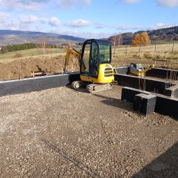 Grzegorz Kozieł - Żółta mini koparka JCB wewnątrz czarnego fundamentu z izolacją, na podsypce żwirowej. W tle pagórkowaty krajobraz i błękitne niebo z chmurami.