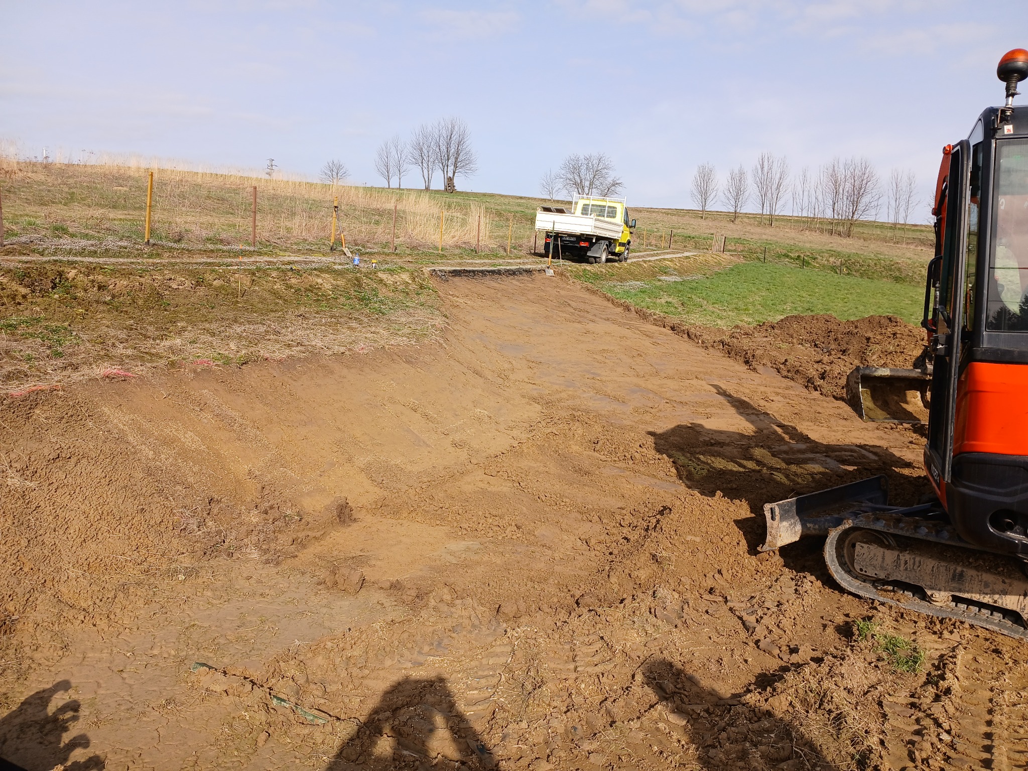 Przygotowany teren pod budowę z widocznym wykopem, małą ciężarówką i koparką w trakcie pracy, z shadowem fotografa w rogu.