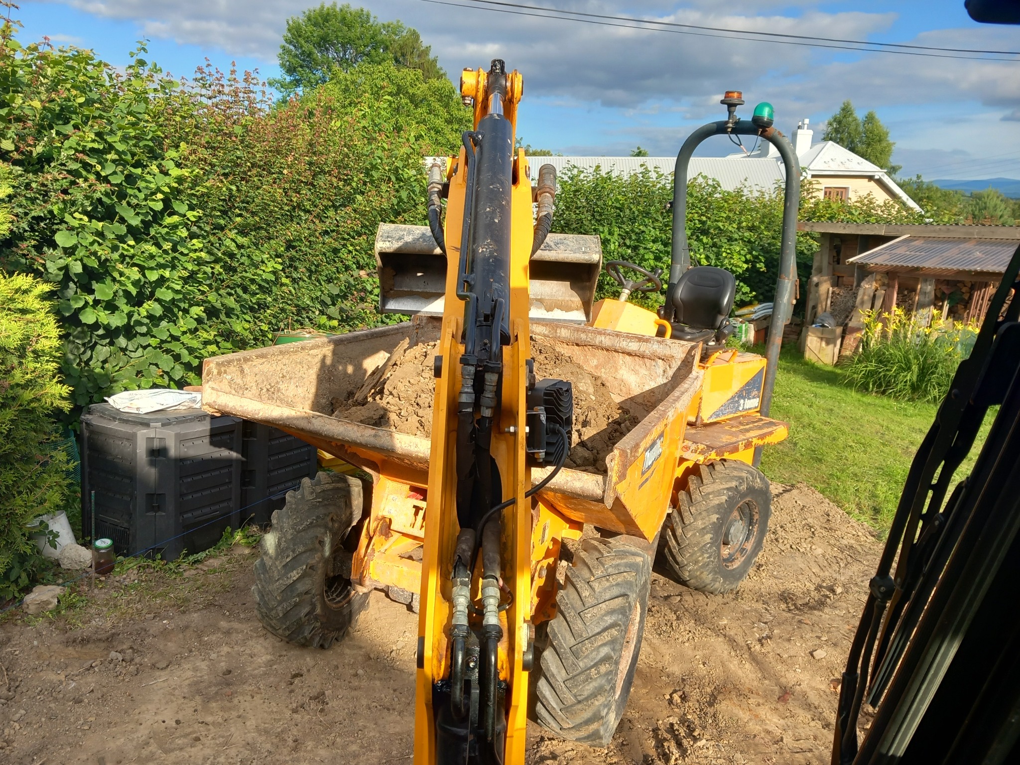 Żółta wywrotka budowlana wypełniona ziemią, widok z perspektywy operatora, w tle zielony żywopłot, dom i szopa.