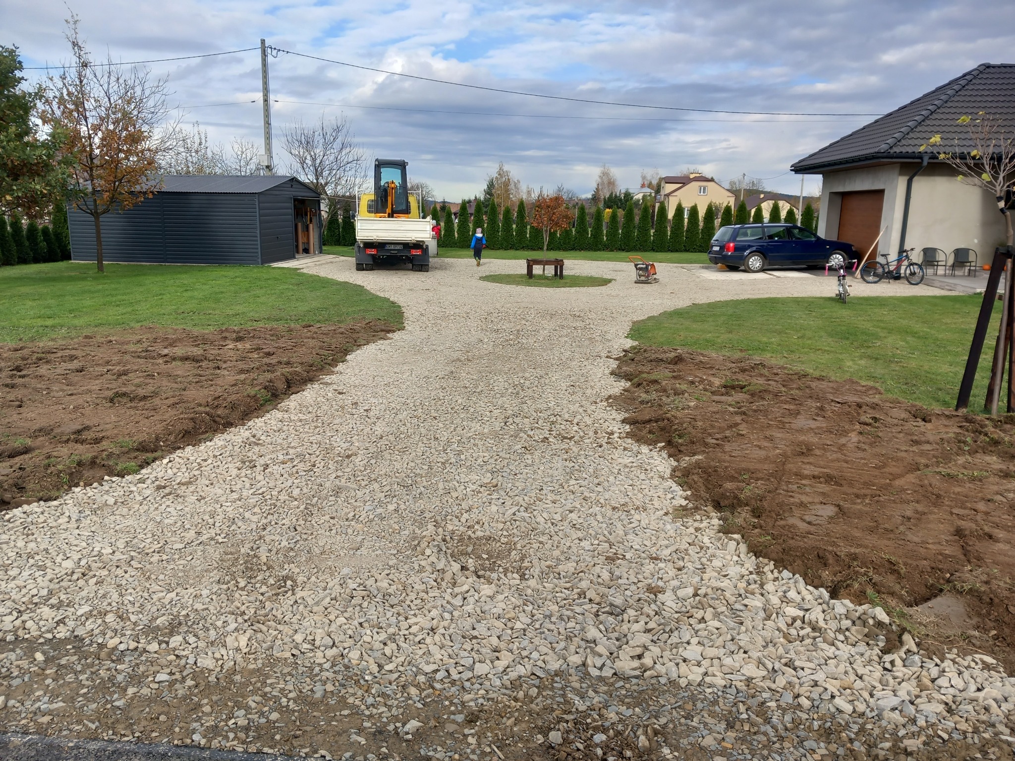 Ukończona żwirowa droga dojazdowa rozwidlająca się przed domem, z widoczną maszyną do zagęszczania gruntu, wywrotką i przygotowanym terenem pod trawnik.