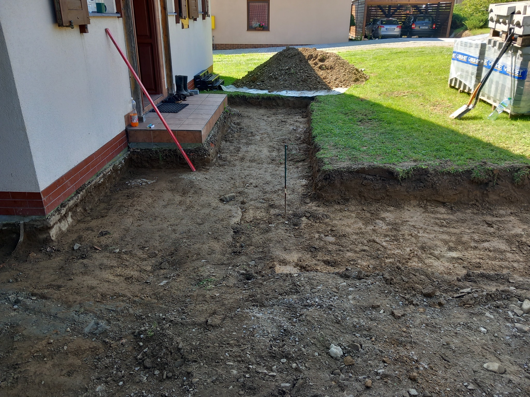 Przygotowany wykop pod kostkę brukową wzdłuż domu, widoczny stos ziemi, paleta kostki brukowej i narzędzia na trawniku
