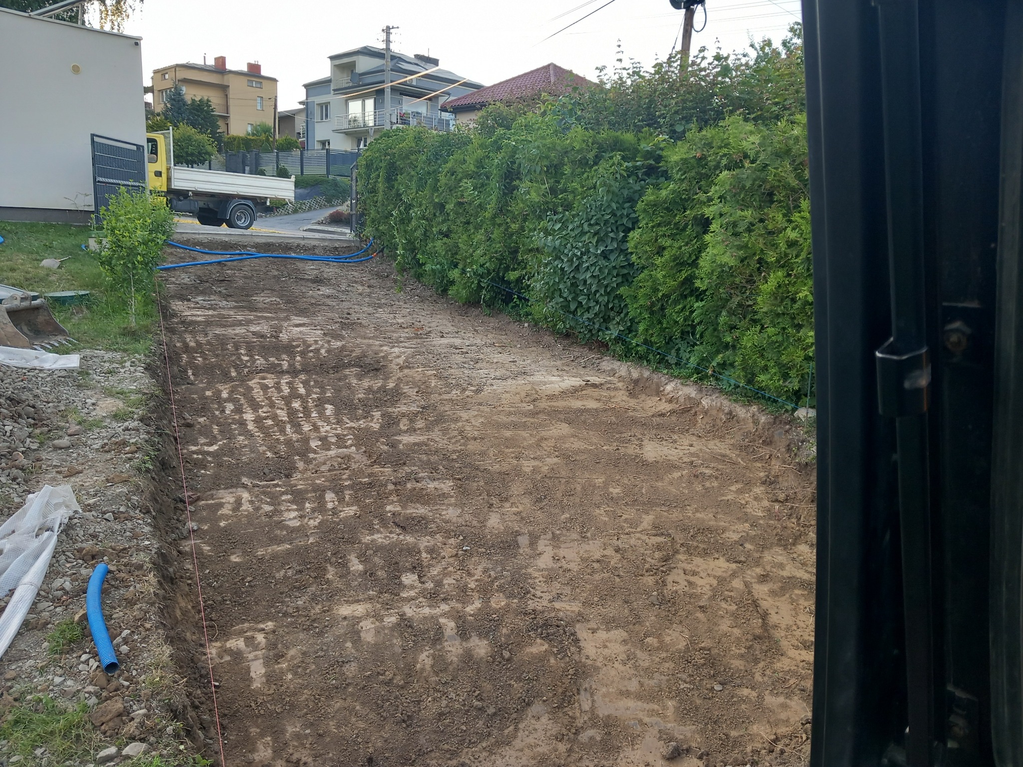 Przygotowany teren pod drogę dojazdową z widocznymi śladami po maszynach, niebieskie rury instalacyjne wzdłuż granicy działki, w tle zaparkowana ciężarówka i zabudowa jednorodzinna.