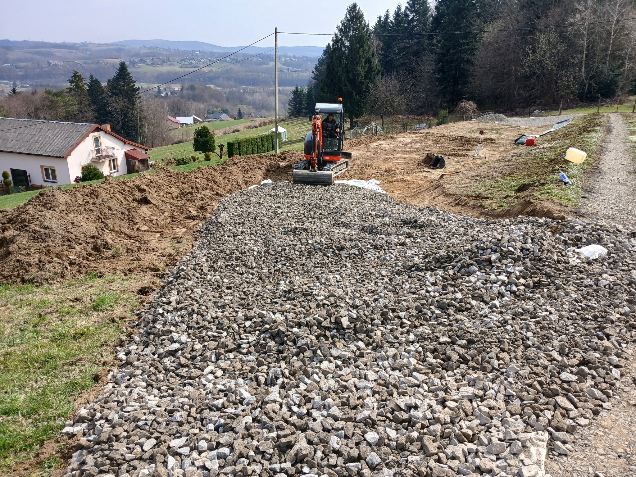 Pomarańczowa mini koparka na terenie budowy z dużą pryzmą kruszywa na pierwszym planie, w tle dom, drzewa i pagórkowaty krajobraz.