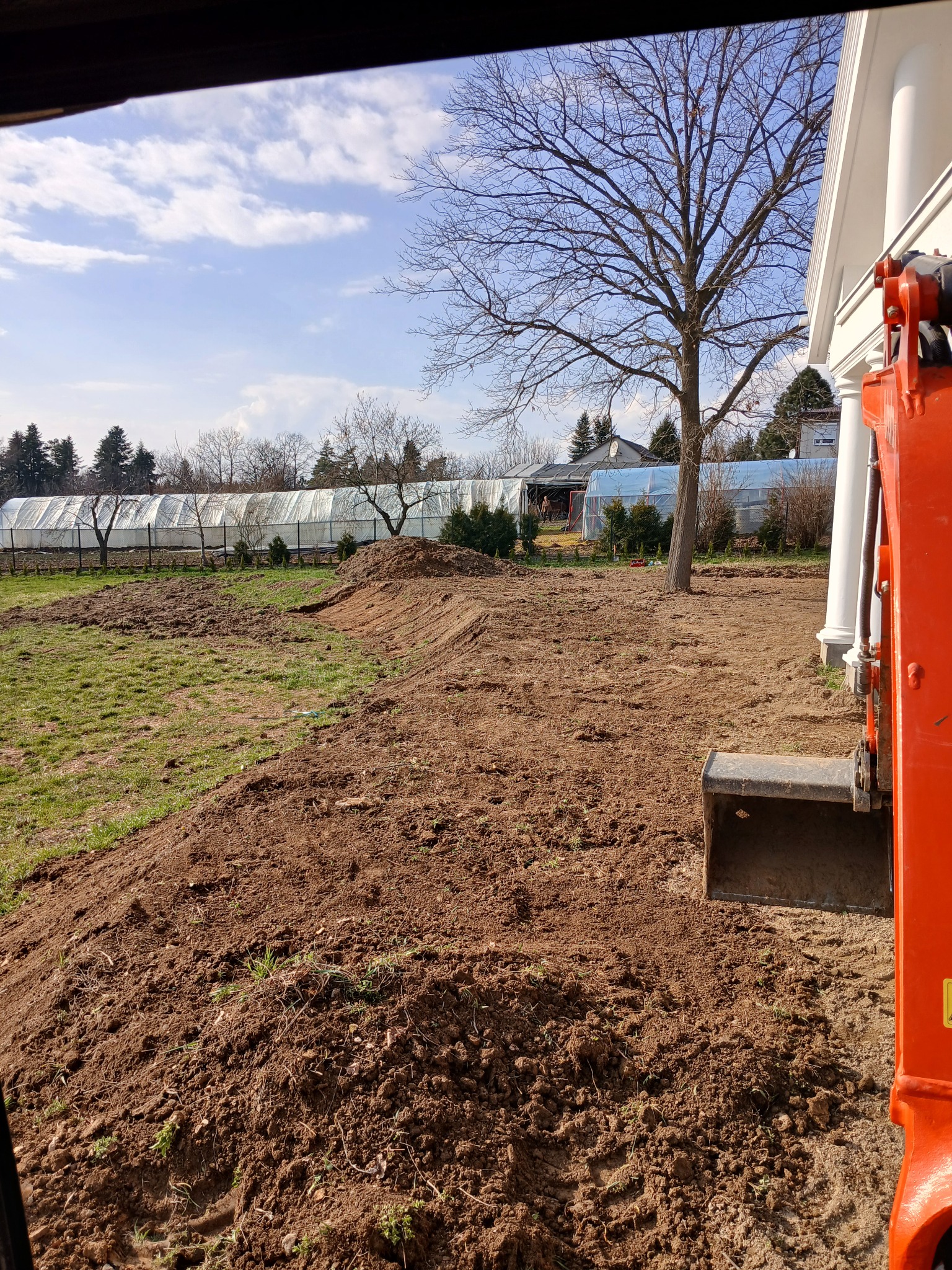 Przygotowanie terenu pod ogród: wyrównana ziemia, widoczny ślad pracy koparki, w tle szklarnie i budynek z kolumnami.