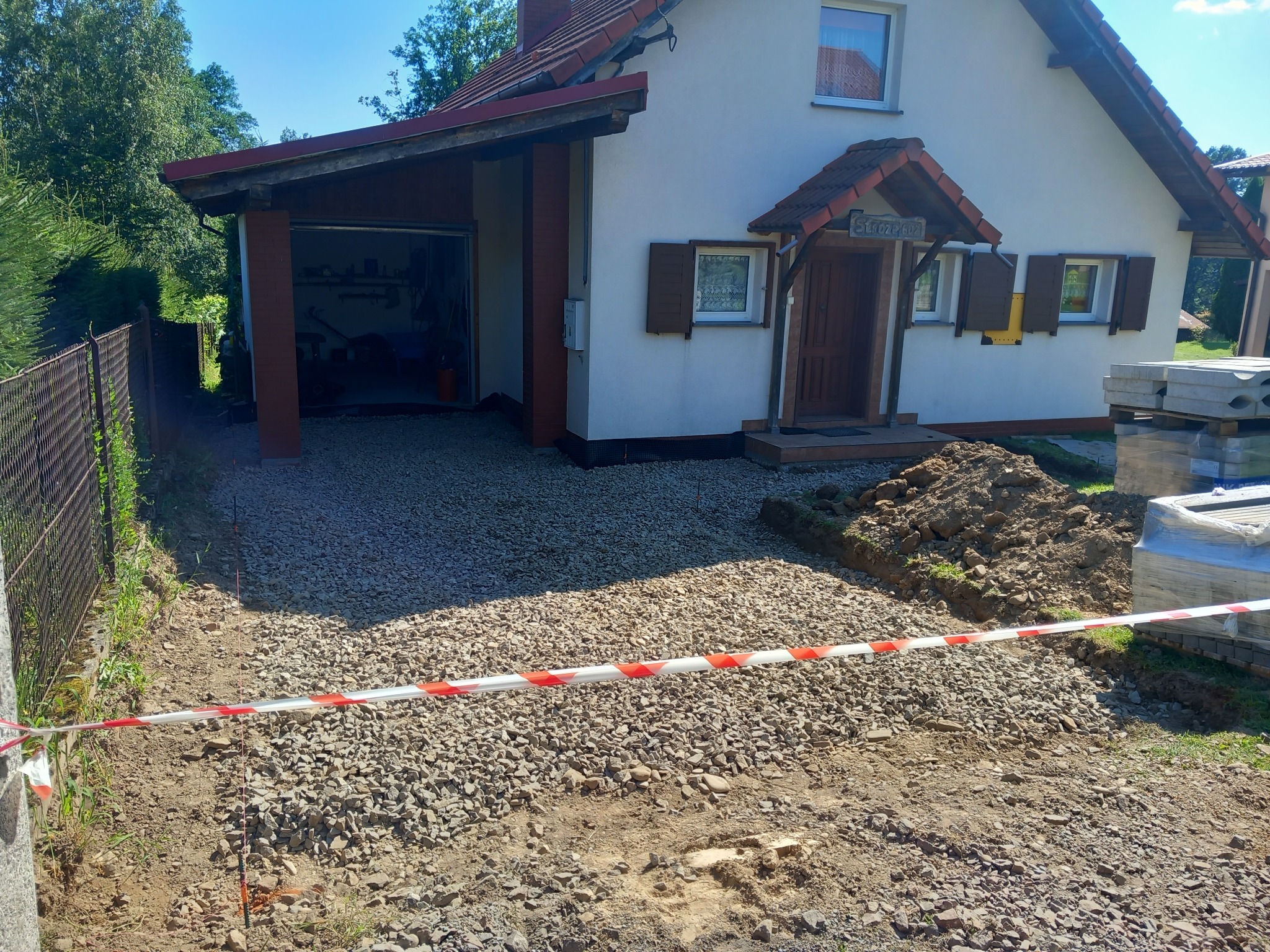 Przygotowanie podłoża z kruszywa przed domem jednorodzinnym z garażem, widoczna taśma ostrzegawcza i paleta z kostką brukową w tle.
