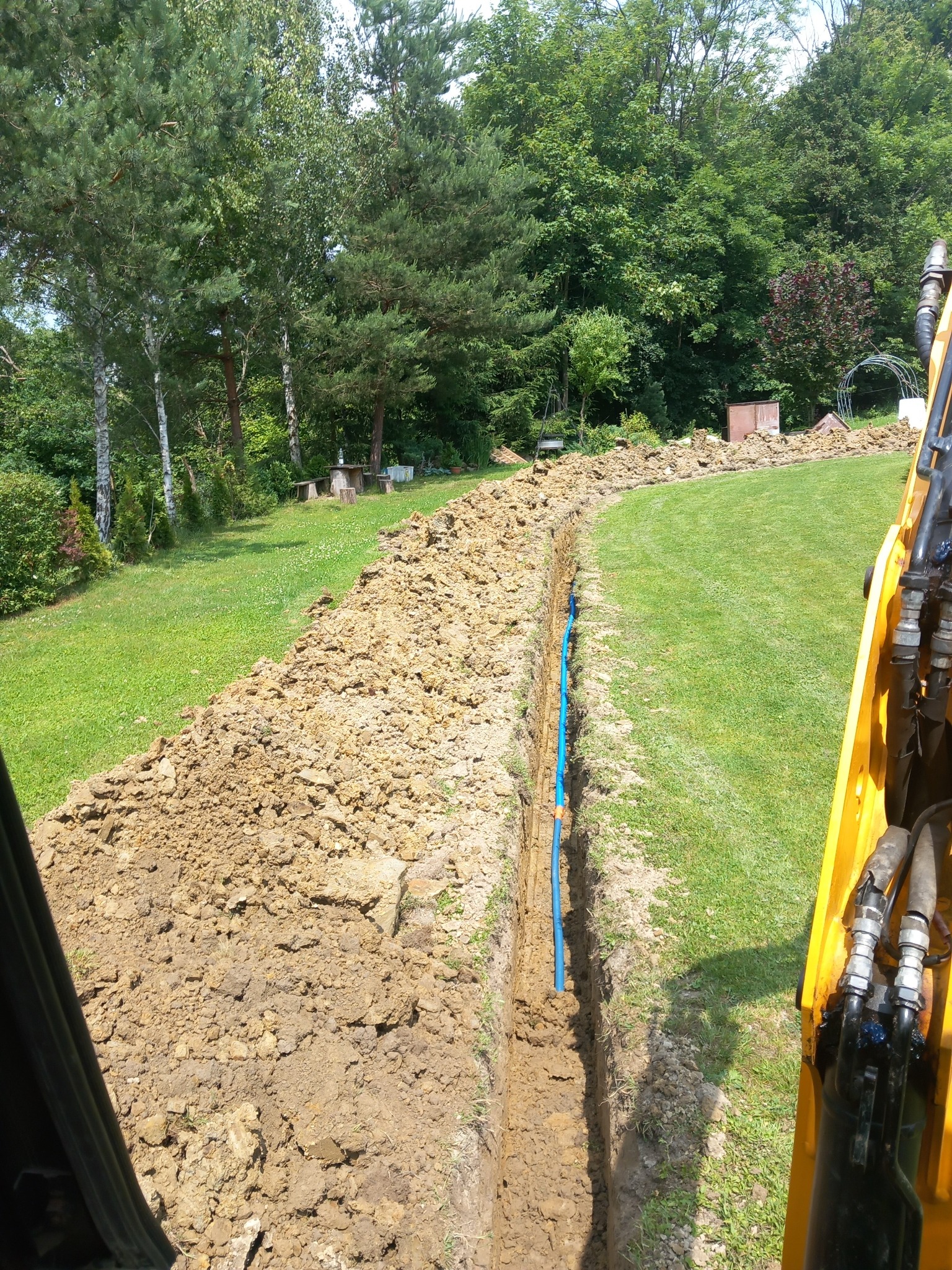 Wąski wykop w trawie, z ułożoną niebieską rurą, widok z kabiny koparki, w tle zieleń drzew.