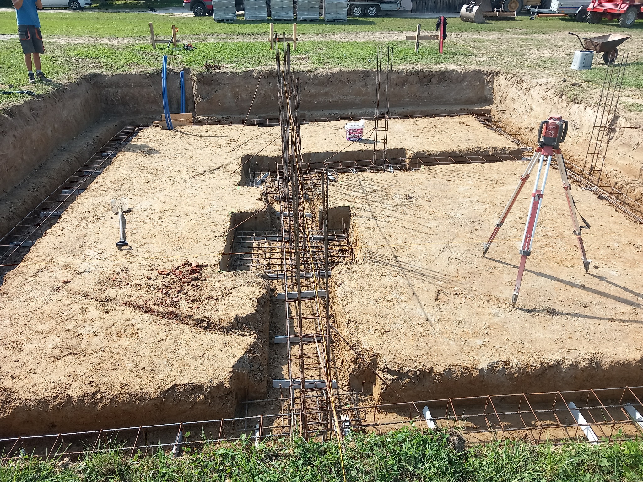 Wykop fundamentowy z ułożonym zbrojeniem i szalunkiem, widoczny niwelator laserowy na trójnogu. Ujęcie z góry, w tle sprzęt budowlany i zieleń.