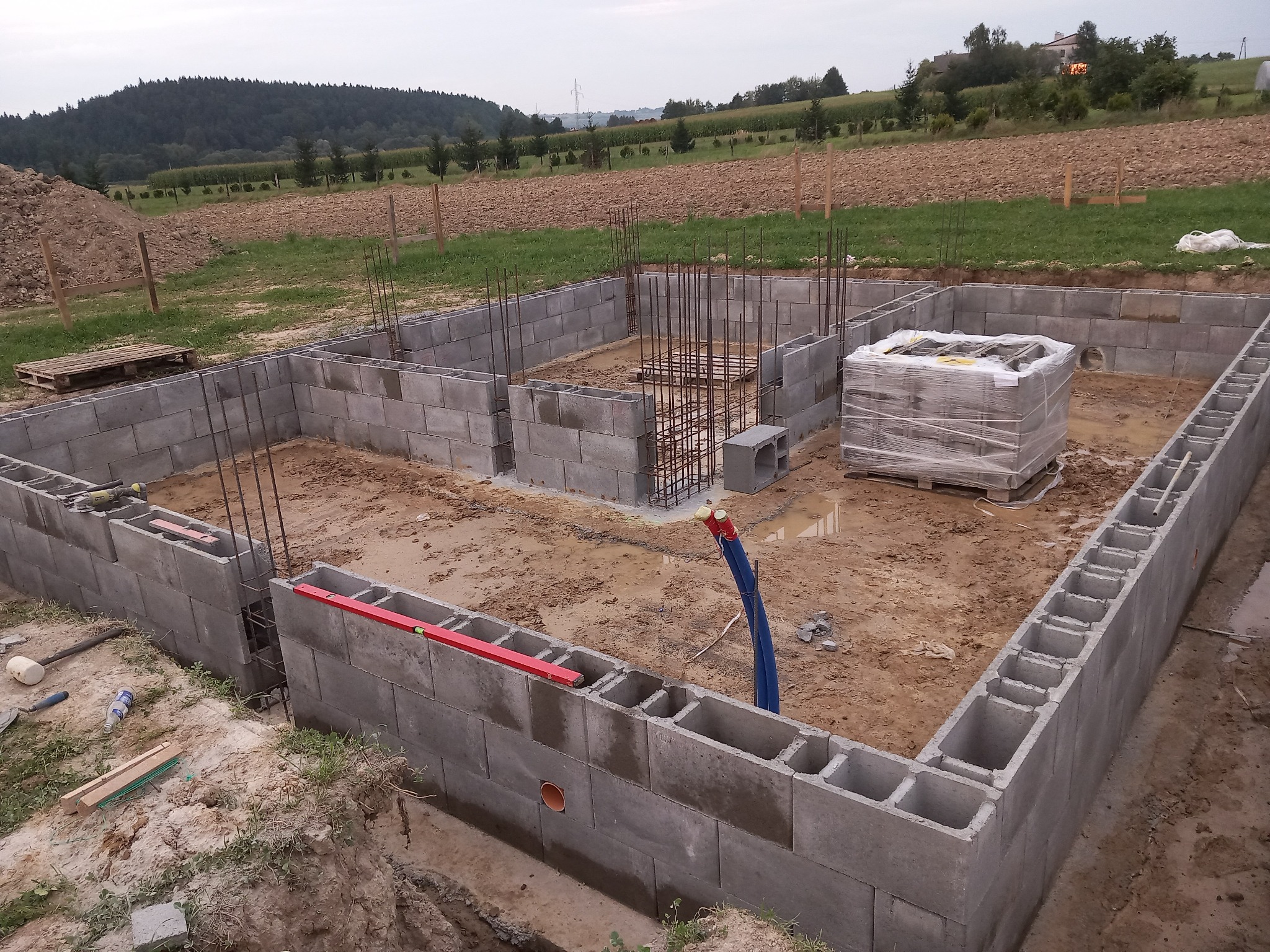 Wznoszenie fundamentów z bloczków betonowych na gruncie. Widoczne zbrojenia, poziomica i paleta bloczków. W tle pola uprawne i las.