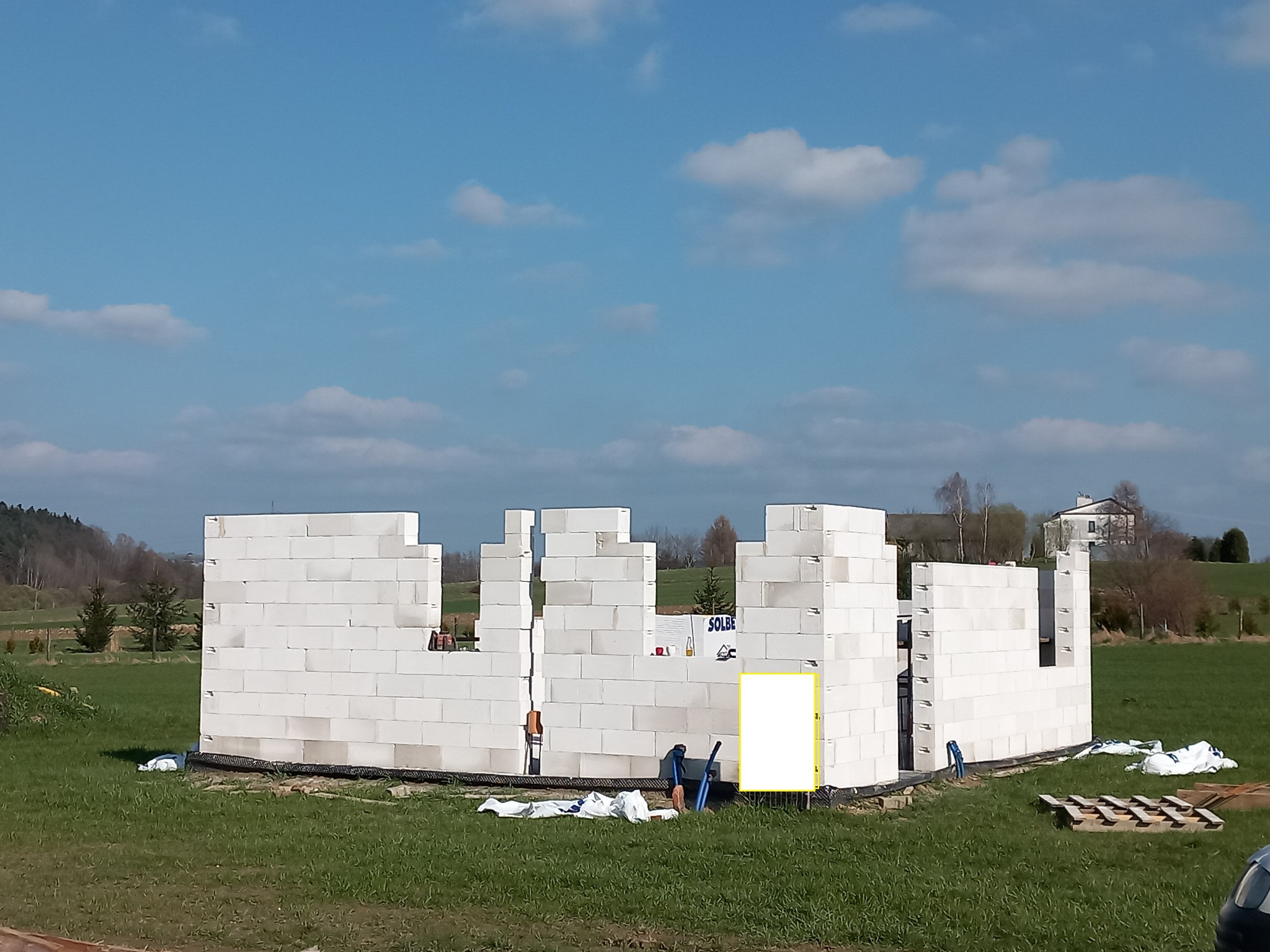 Wznoszenie ścian parteru domu z białych bloczków, widok z zewnątrz na tle zielonej łąki i błękitnego nieba z chmurami. Widoczne narzędzia i materiały budowlane.