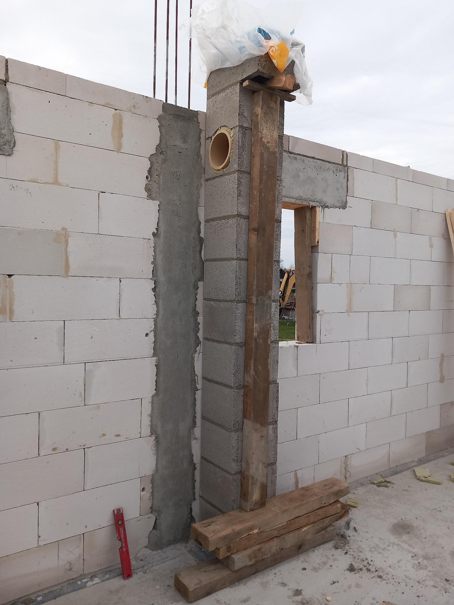 Ściana z betonu komórkowego w trakcie budowy, z widocznym otworem okiennym i elementami szalunku. Na ścianie widoczne ślady zaprawy i poziomica.