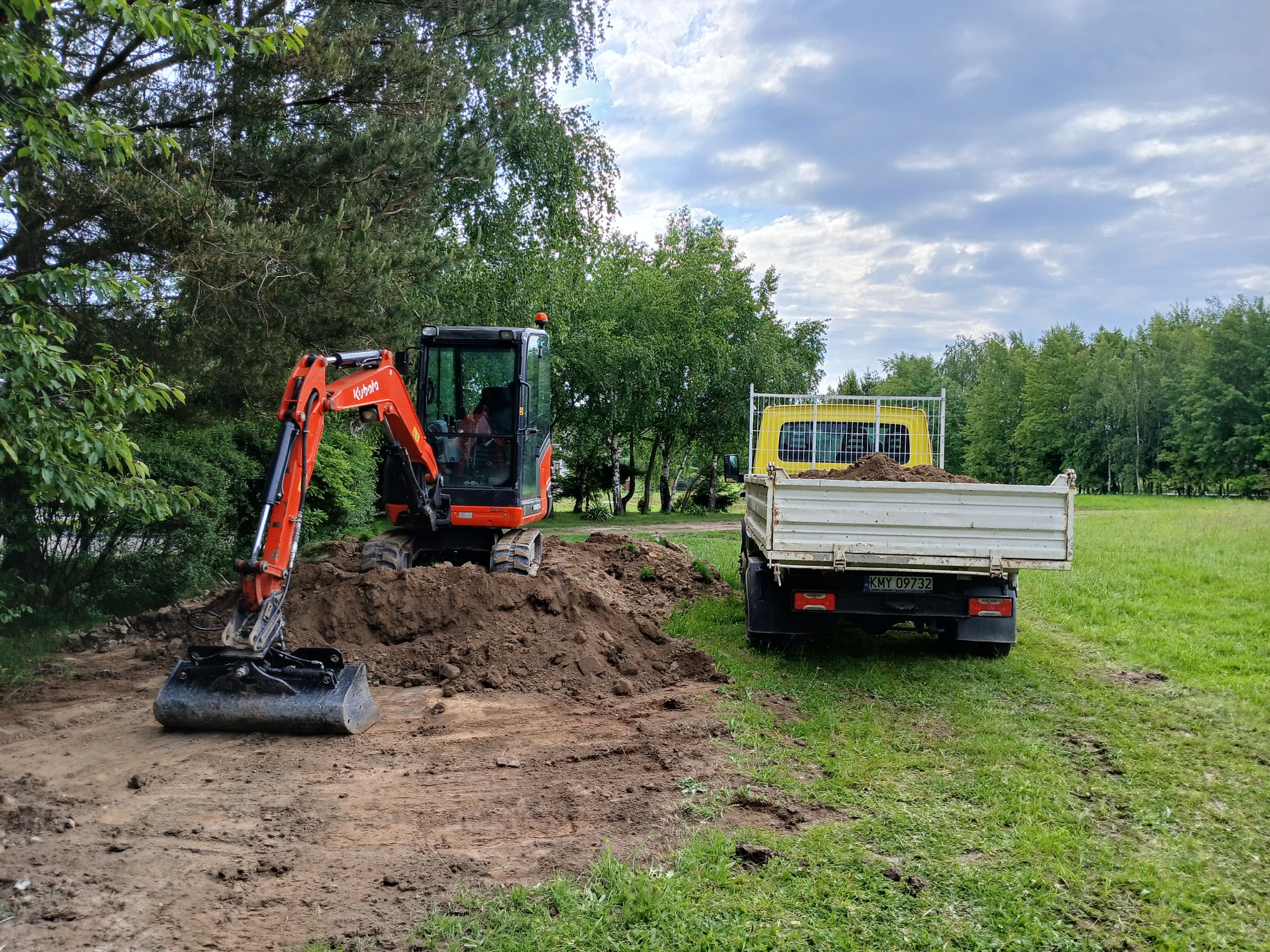 Pomarańczowa minikoparka Kubota i żółta wywrotka na trawiastym terenie podczas prac ziemnych. Widoczne ślady opon i świeżo usypana ziemia.
