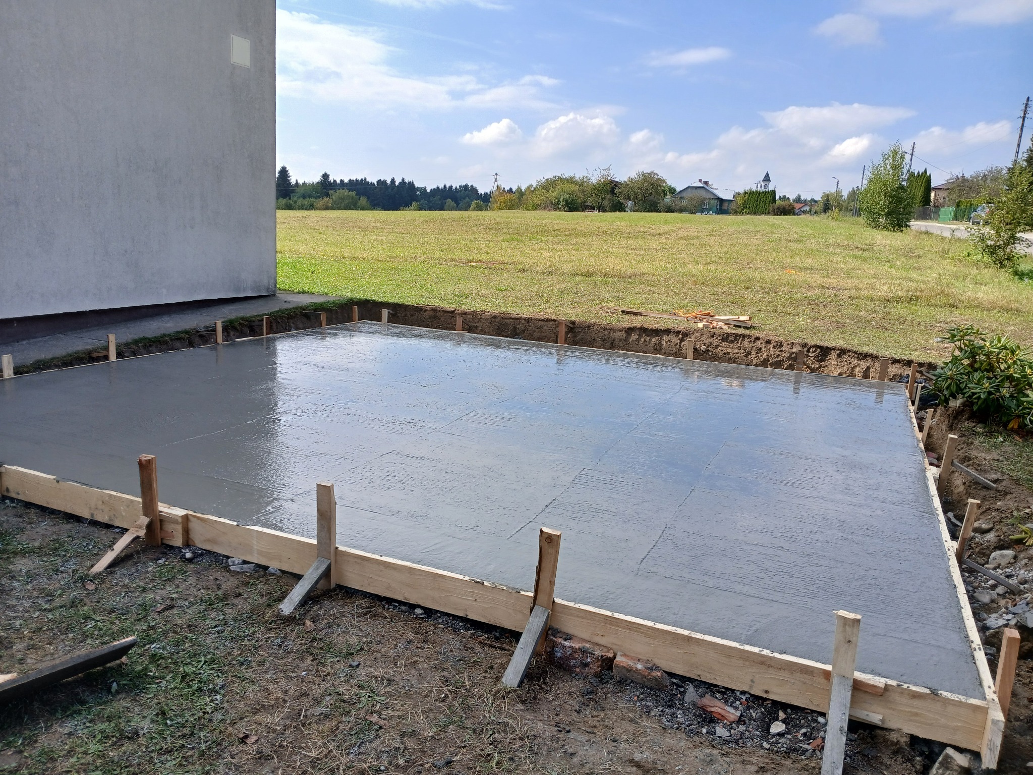 Świeżo wylany, gładki fundament pod budynek z drewnianym szalunkiem, na tle zielonego pola i błękitnego nieba z chmurami. Widok z poziomu gruntu.