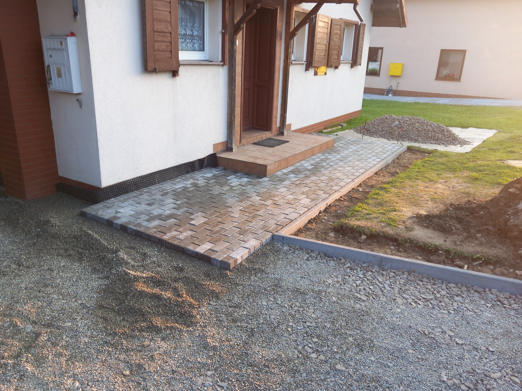 Ułożona kostka brukowa prowadząca do wejścia domu, obok widoczna warstwa podsypki i obrzeża. Częściowo widoczny budynek z brązowymi okiennicami.