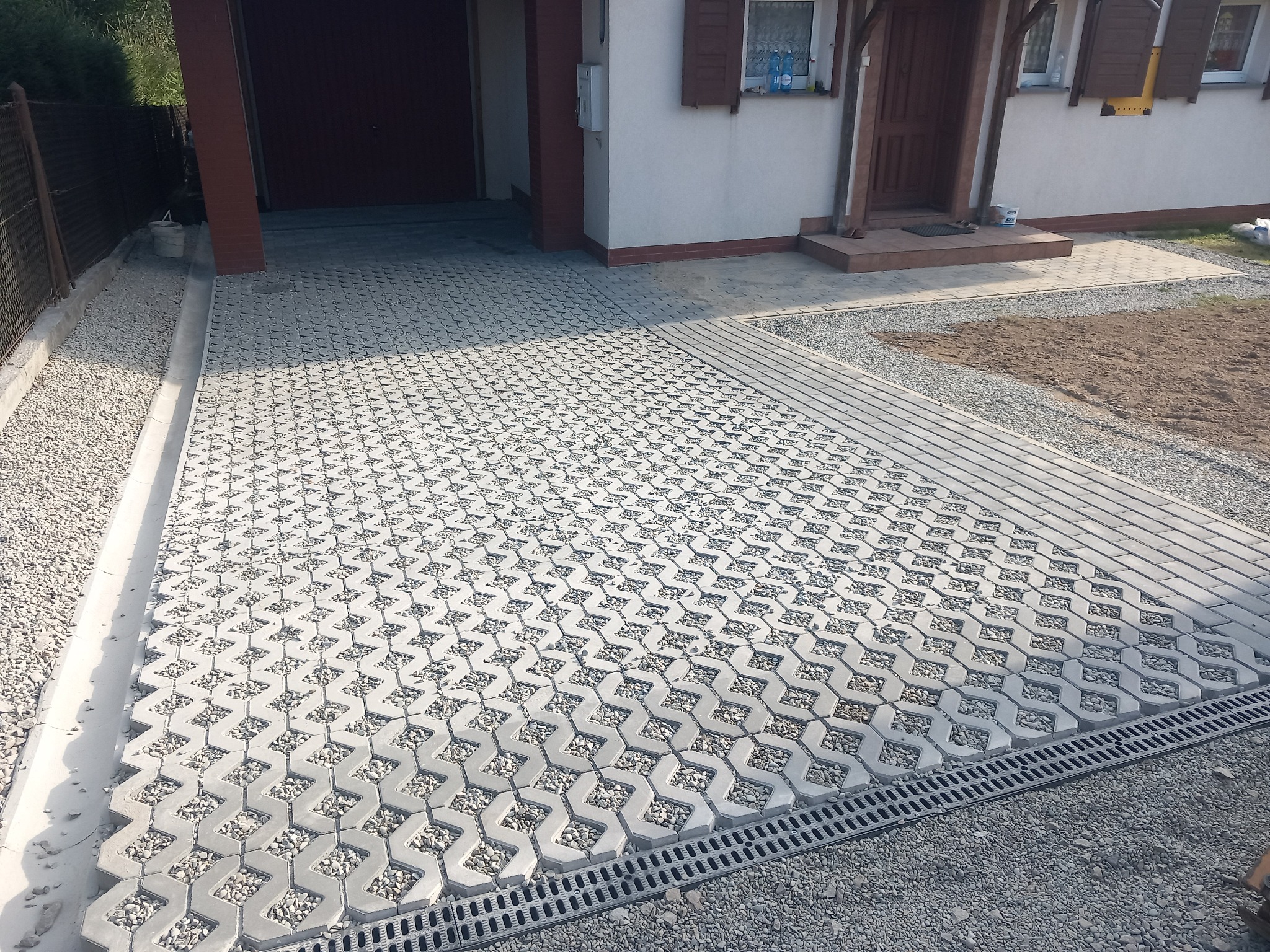 Ułożona szara ażurowa kostka brukowa z wypełnieniem żwirowym na podjeździe przed domem. Widoczny system odprowadzania wody i fragment trawnika.